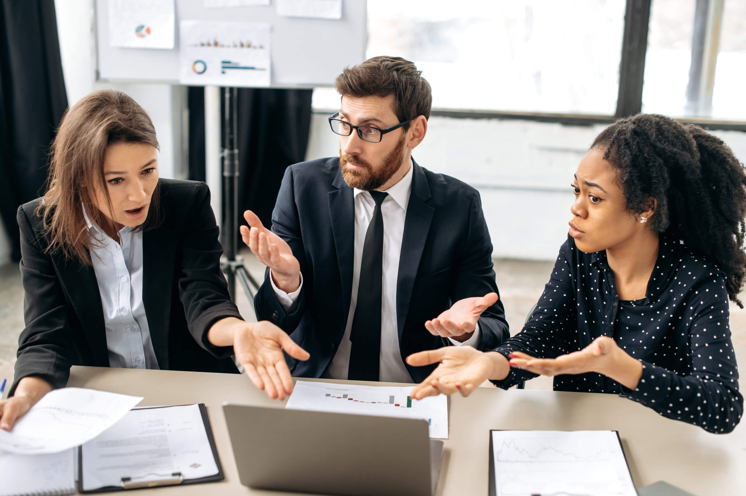 Three professionals sit at a table, looking surprised and confused as they discuss something in front of a laptop. Papers with charts are spread on the table. The background shows a whiteboard with graphs.