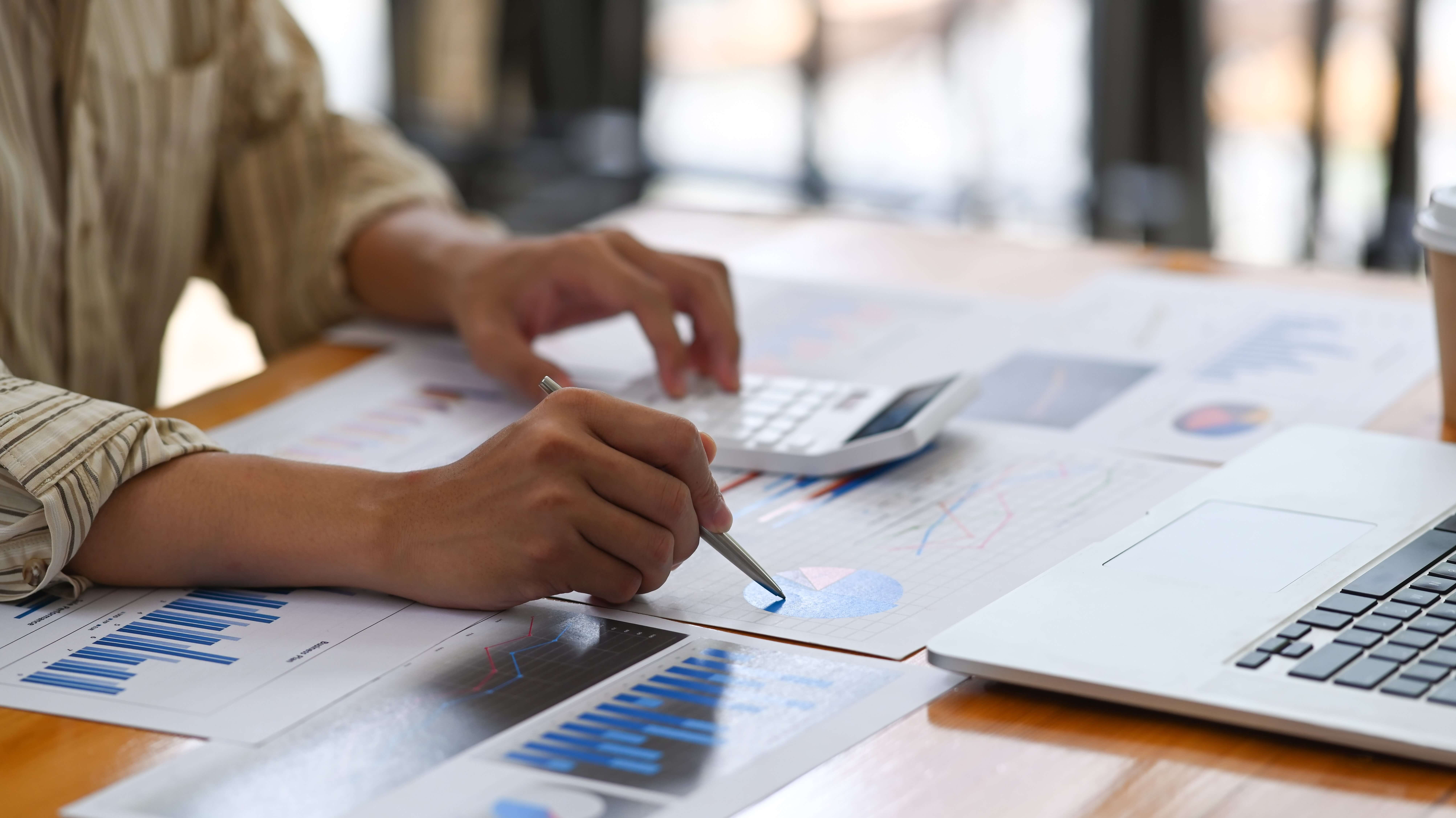 Person using a calculator and pointing at financial charts on a table, with a laptop nearby. The focus is on the charts and data analysis, indicating a work or study environment related to finance or business.