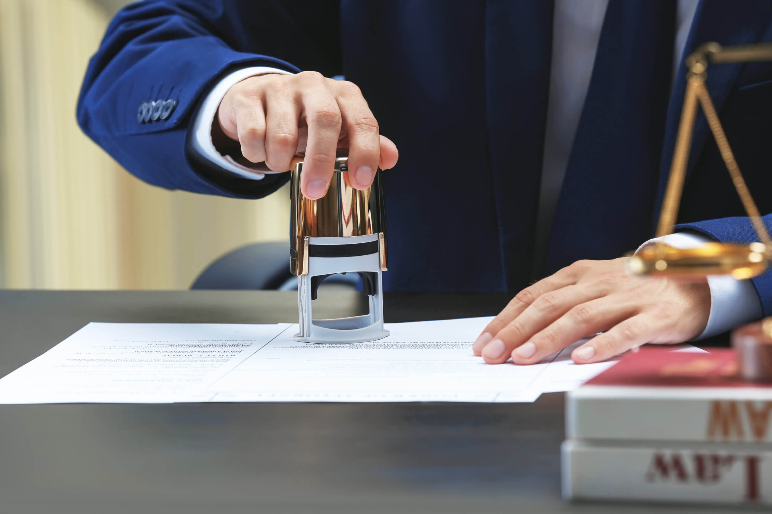 A person in a suit stamps a document on a desk. Nearby are a stack of books and a small golden justice scale. The setting appears to be an office or legal environment.