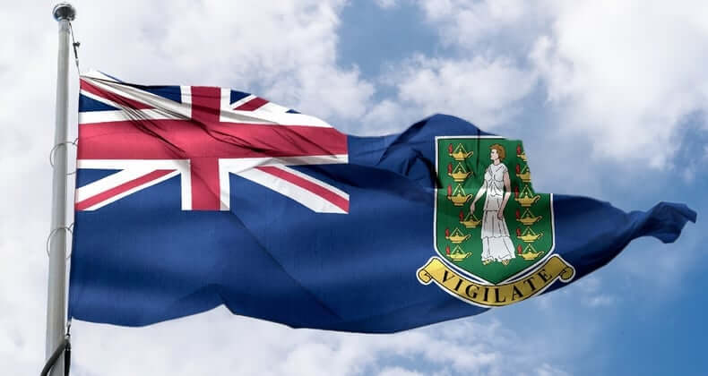 The British Virgin Islands flag features a Union Jack in the canton and the territory's coat of arms on a blue field. The coat of arms displays Saint Ursula holding a gold lamp, with the motto "Vigilate" below. The flag is waving against a cloudy sky.