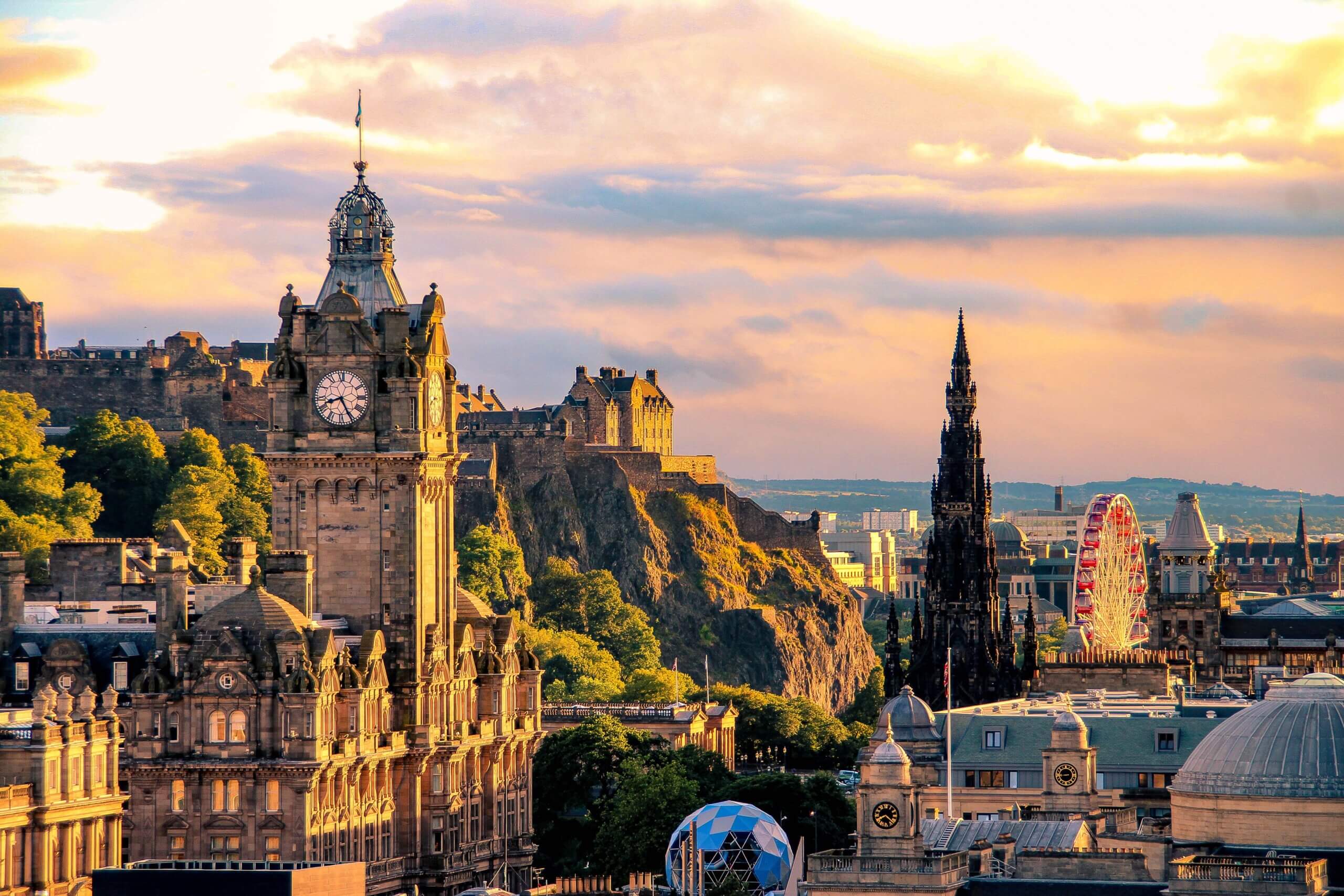 A scenic view of Edinburgh at sunset, showcasing the historic Balmoral Hotel clock tower, Edinburgh Castle on a hill, the Scott Monument, and a Ferris wheel. The sky is lit with warm hues of orange and yellow, adding a glow to the city's architecture.