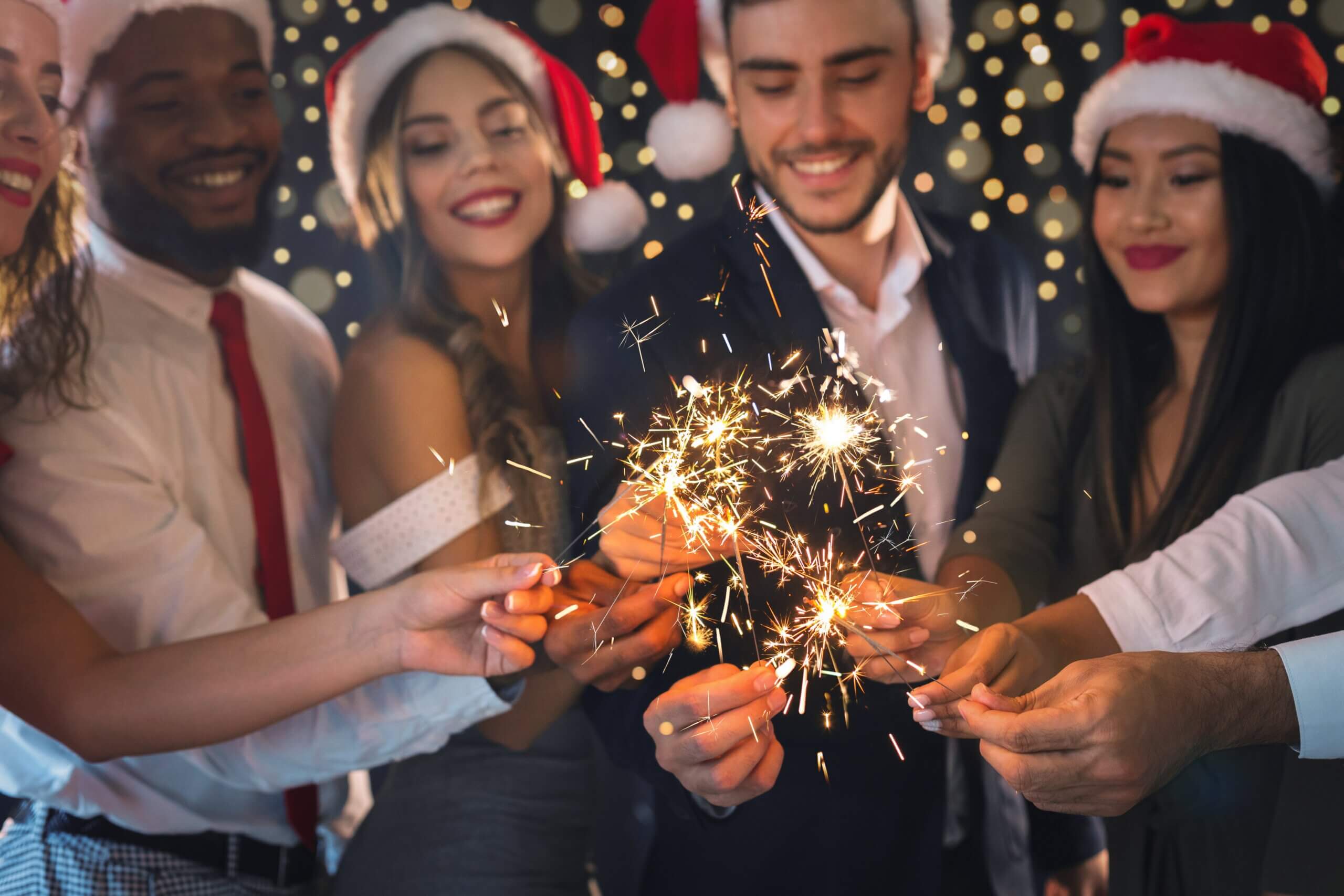 A group of people wearing festive clothing and Santa hats is gathered closely together, holding sparklers. They are smiling and celebrating against a background of twinkling lights, creating a joyful and festive atmosphere.