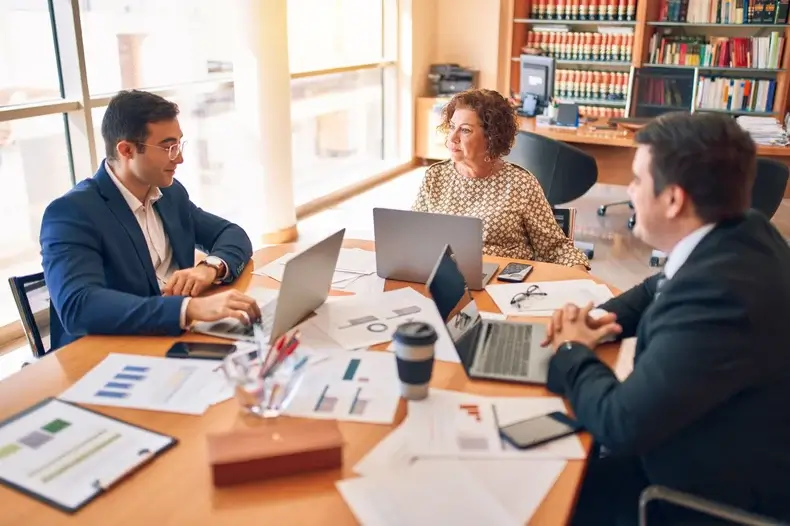 Three people sit around a conference table with laptops, discussing business. Documents and charts are spread out. A window with natural light is on the left, and bookshelves are in the background. The atmosphere is collaborative and professional.