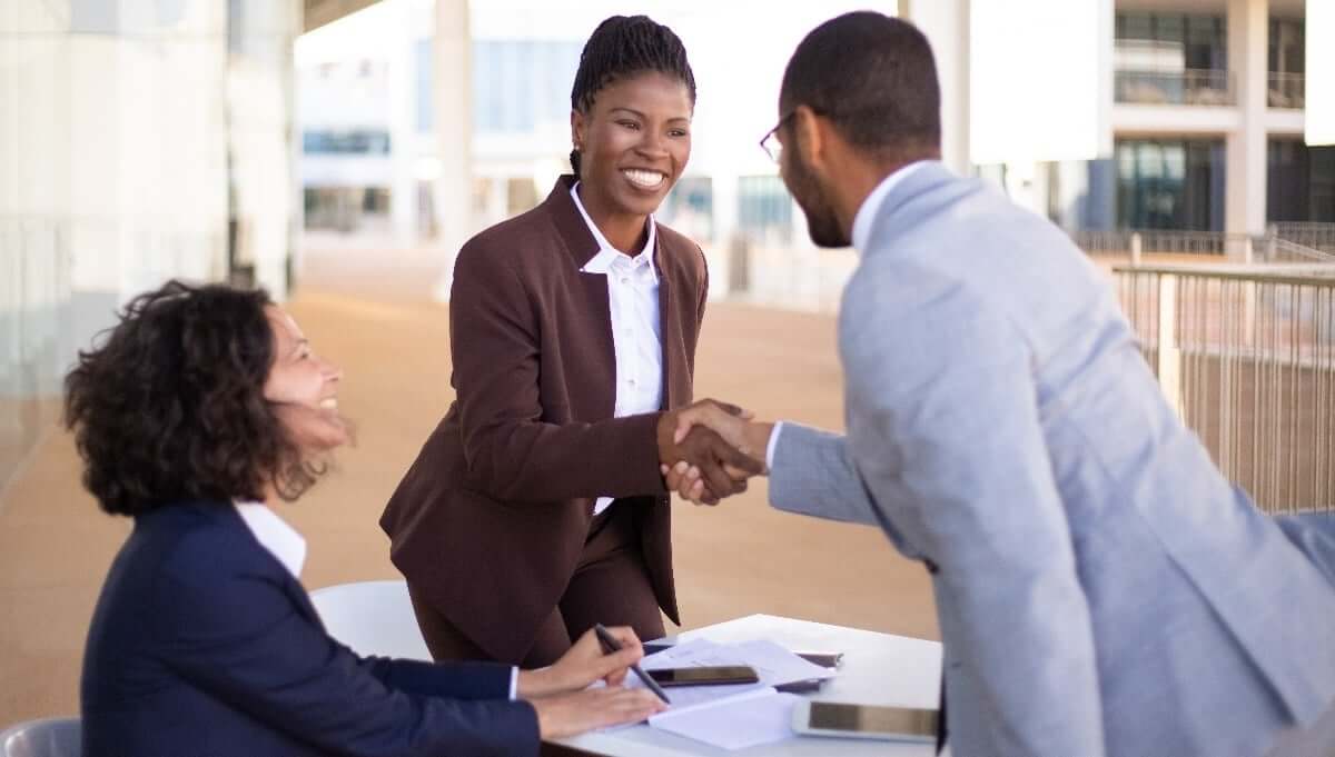 Three professionals outdoors: two women sitting at a table with documents, one smiling and the other with curly hair. A man in a suit is standing, shaking hands with the smiling woman. Modern building in the background.