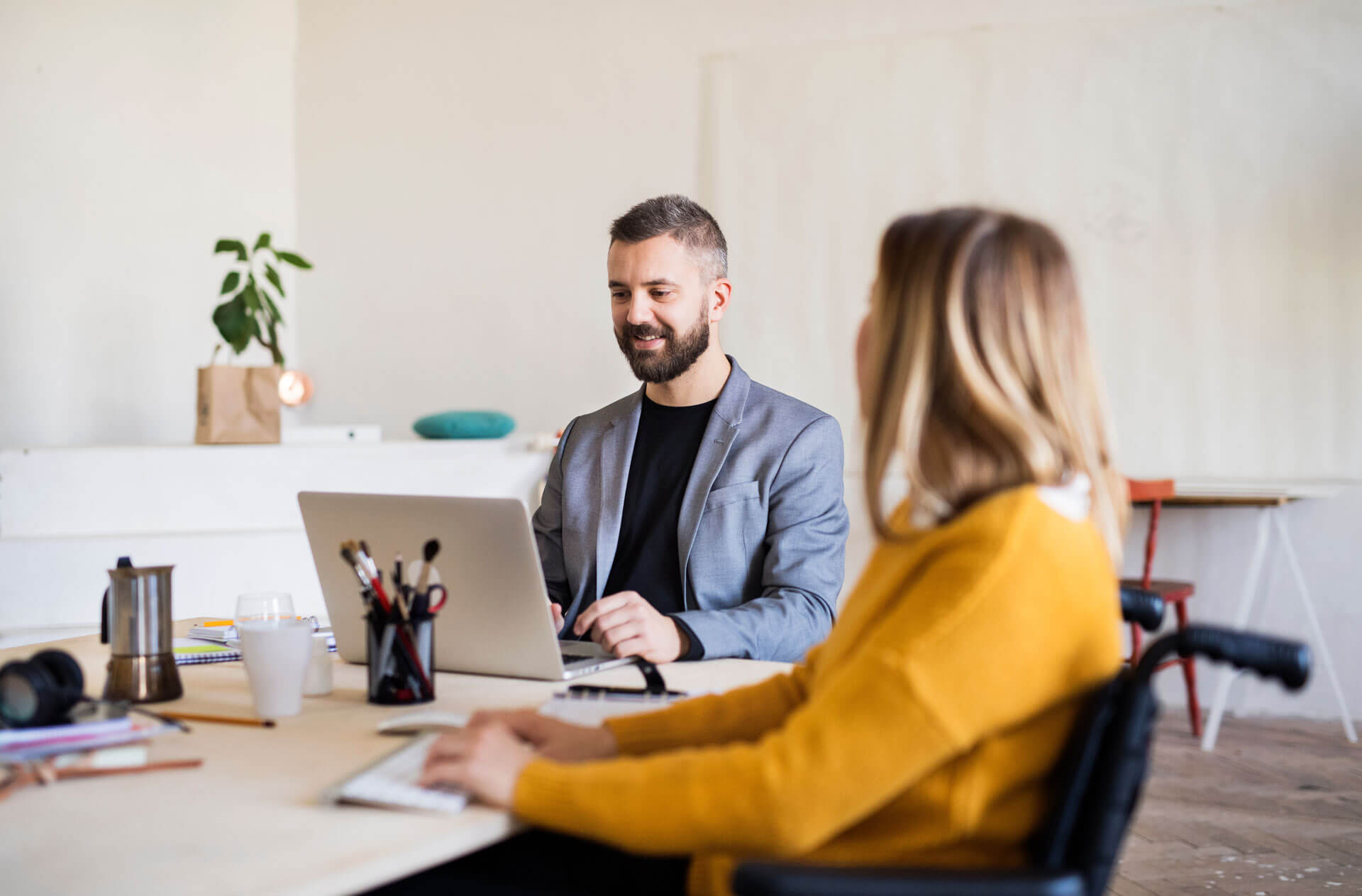 Two people sitting at a table in an office setting. One is a man wearing a gray blazer working on a laptop. The other is a woman with long hair, wearing a mustard sweater, sitting in a wheelchair. A plant and office supplies are visible on the table.