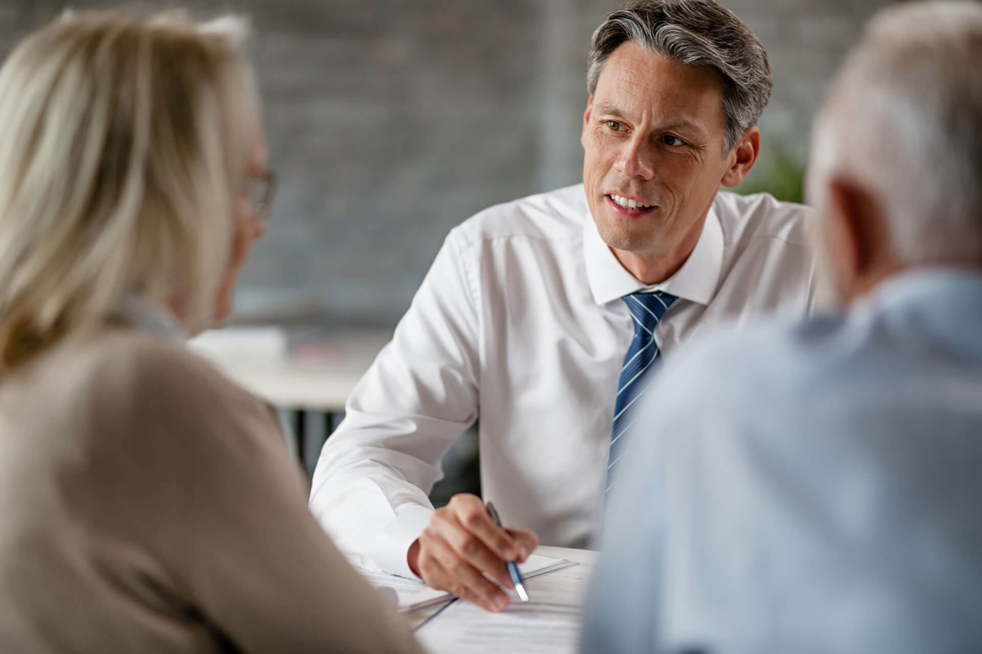 A man in a white shirt and tie sits at a table, talking to two people who are partially visible. He is holding a pen and looking engaged in the conversation. The setting appears to be an office or professional environment.