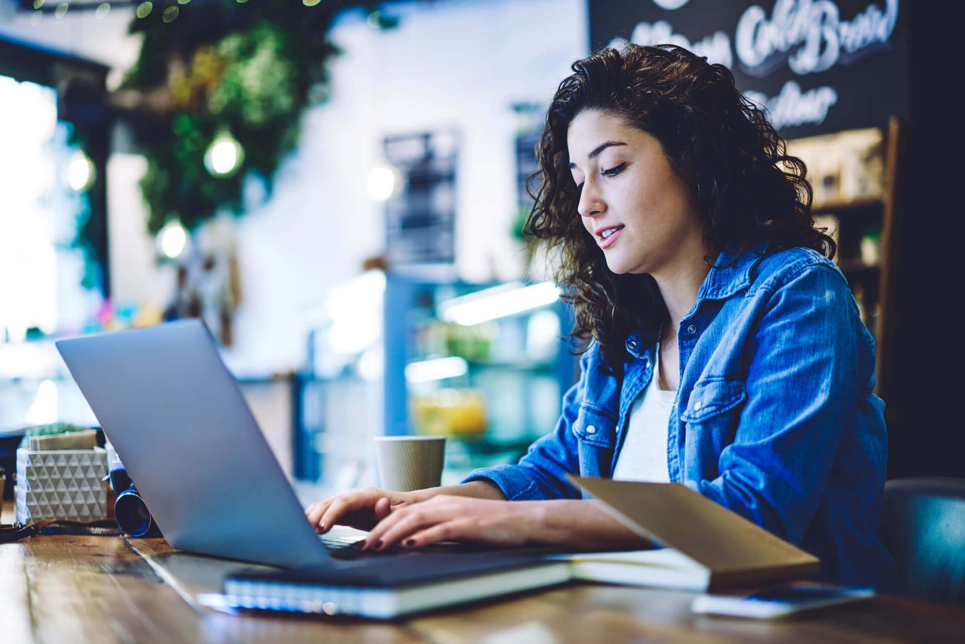 A woman with curly hair wearing a denim shirt is sitting at a wooden table in a cozy café, typing on a laptop. An open book lies next to her, and there is a coffee cup on the table. The background features greenery and soft lighting.