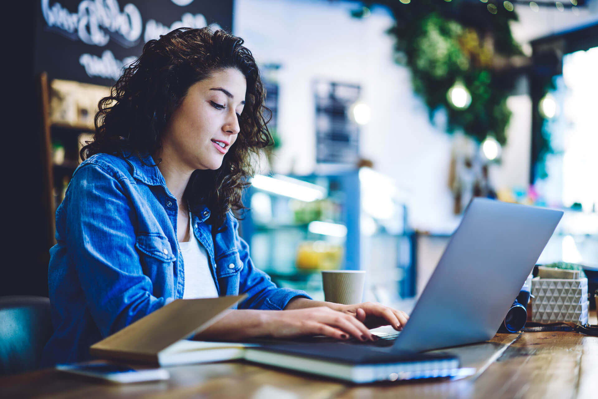 A woman with curly hair wearing a denim shirt is sitting at a wooden table in a cozy café, typing on a laptop. An open book lies next to her, and there is a coffee cup on the table. The background features greenery and soft lighting.