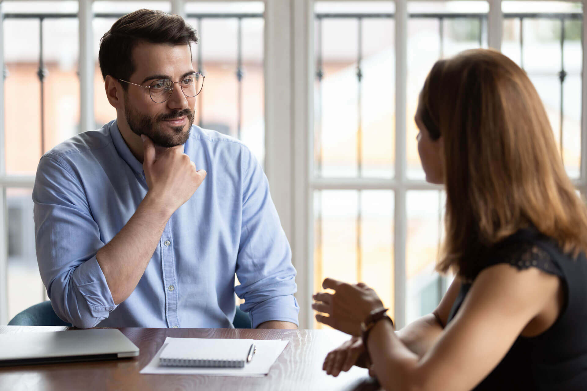 A man and a woman sit across from each other at a table, engaged in a conversation. The man, wearing glasses and a light blue shirt, listens attentively with his hand on his chin. The woman, seen from behind, gestures with her hands. A notebook and pen are on the table.