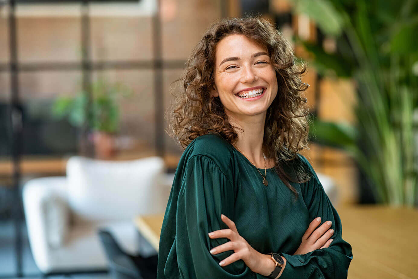 A smiling woman with curly hair wearing a dark green shirt stands with crossed arms in a modern, well-lit room with indoor plants, representing Cogency Global’s simple change of registered agent services. This image links to a video on how to change your registered agent with Cogency Global when clicked.