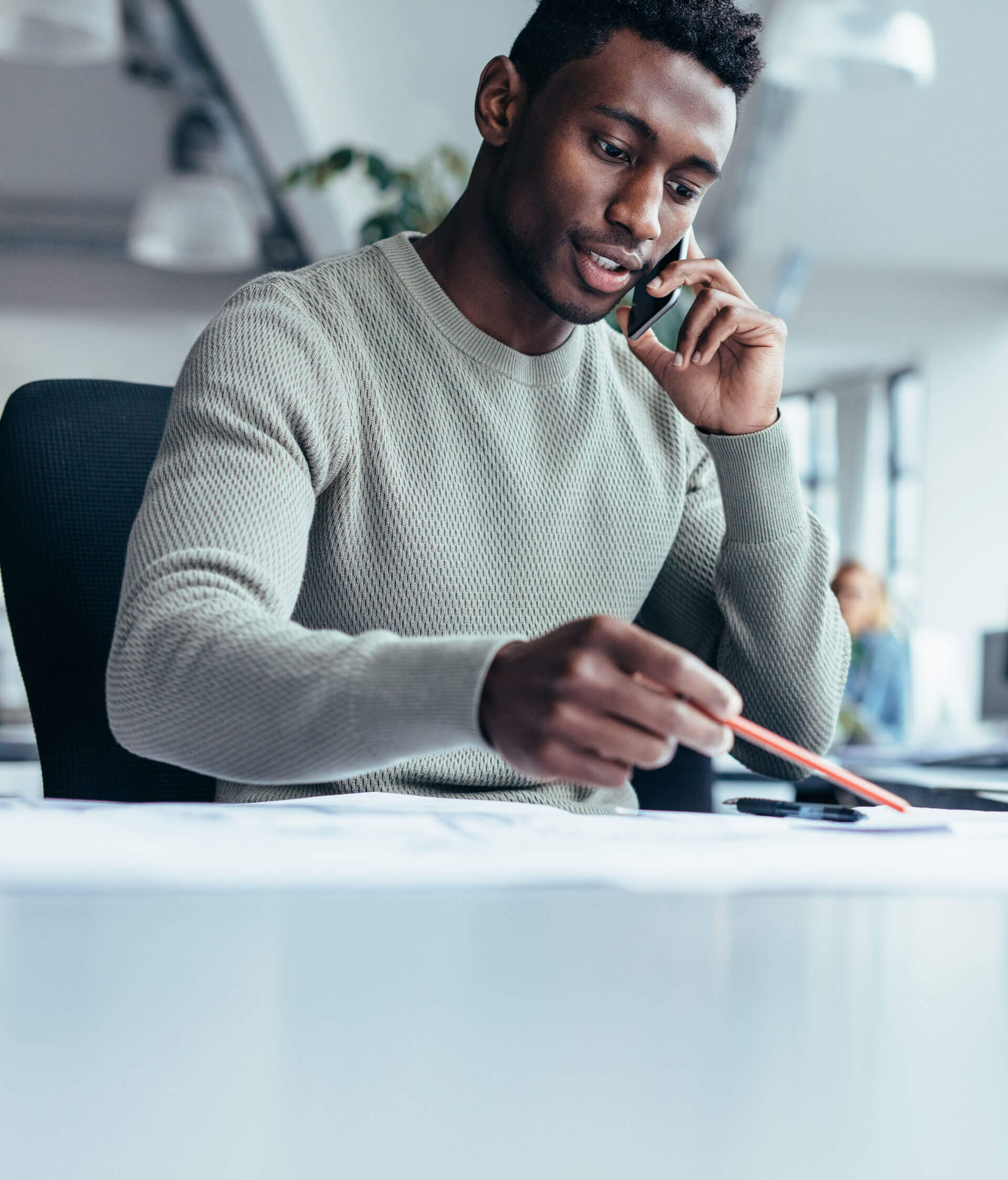 Man in a sweater sits at a desk, talking on a phone and pointing at documents with a red pencil. Office setting with blurred background and a person working at another desk.