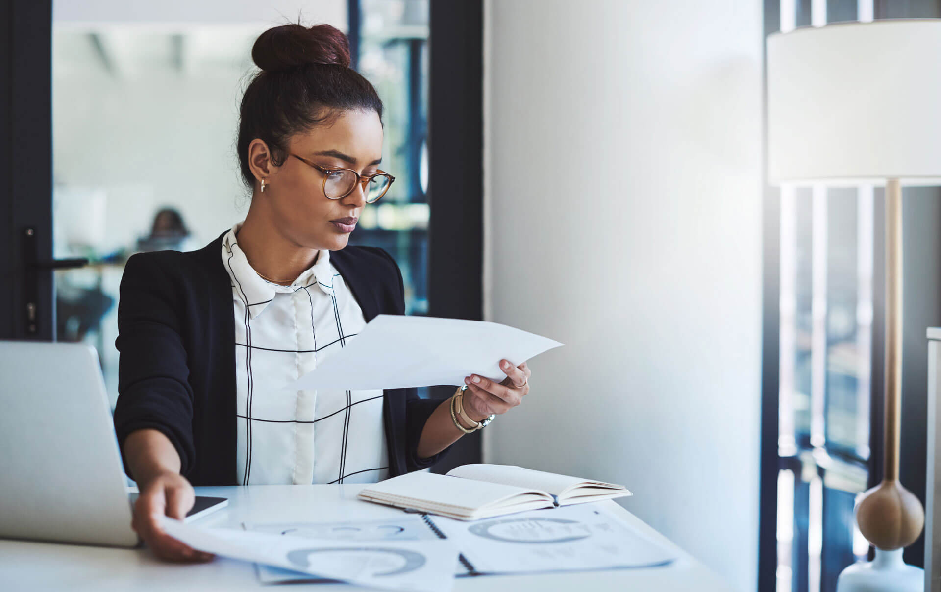 A person with glasses and a bun hairstyle sits at a desk, looking at papers. They are wearing a black blazer over a checkered shirt. A laptop is open on the desk, along with more papers and a notebook. Natural light fills the modern office space.