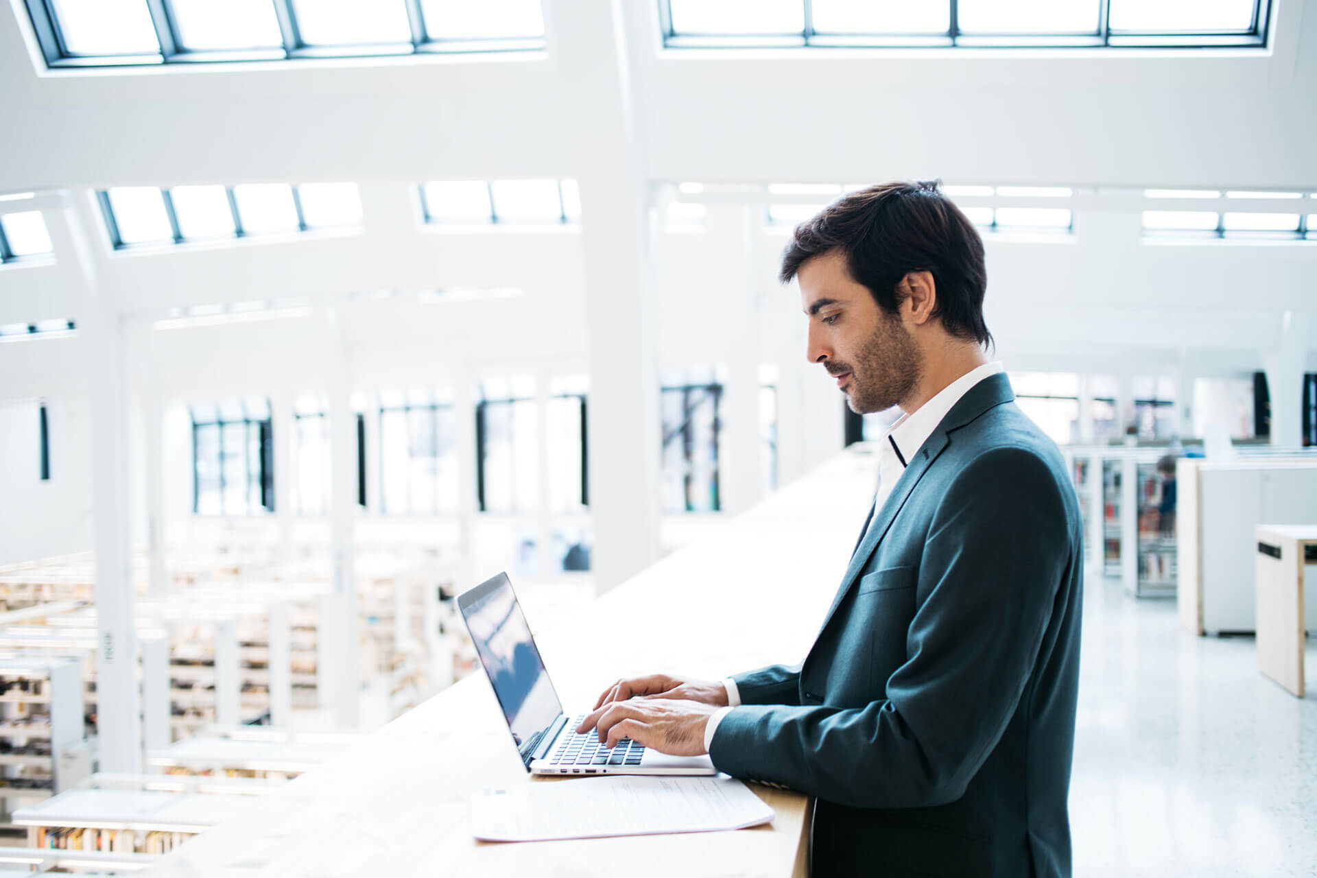 A man in a business suit stands and types on a laptop in a bright, modern office space with large windows and geometric design elements.