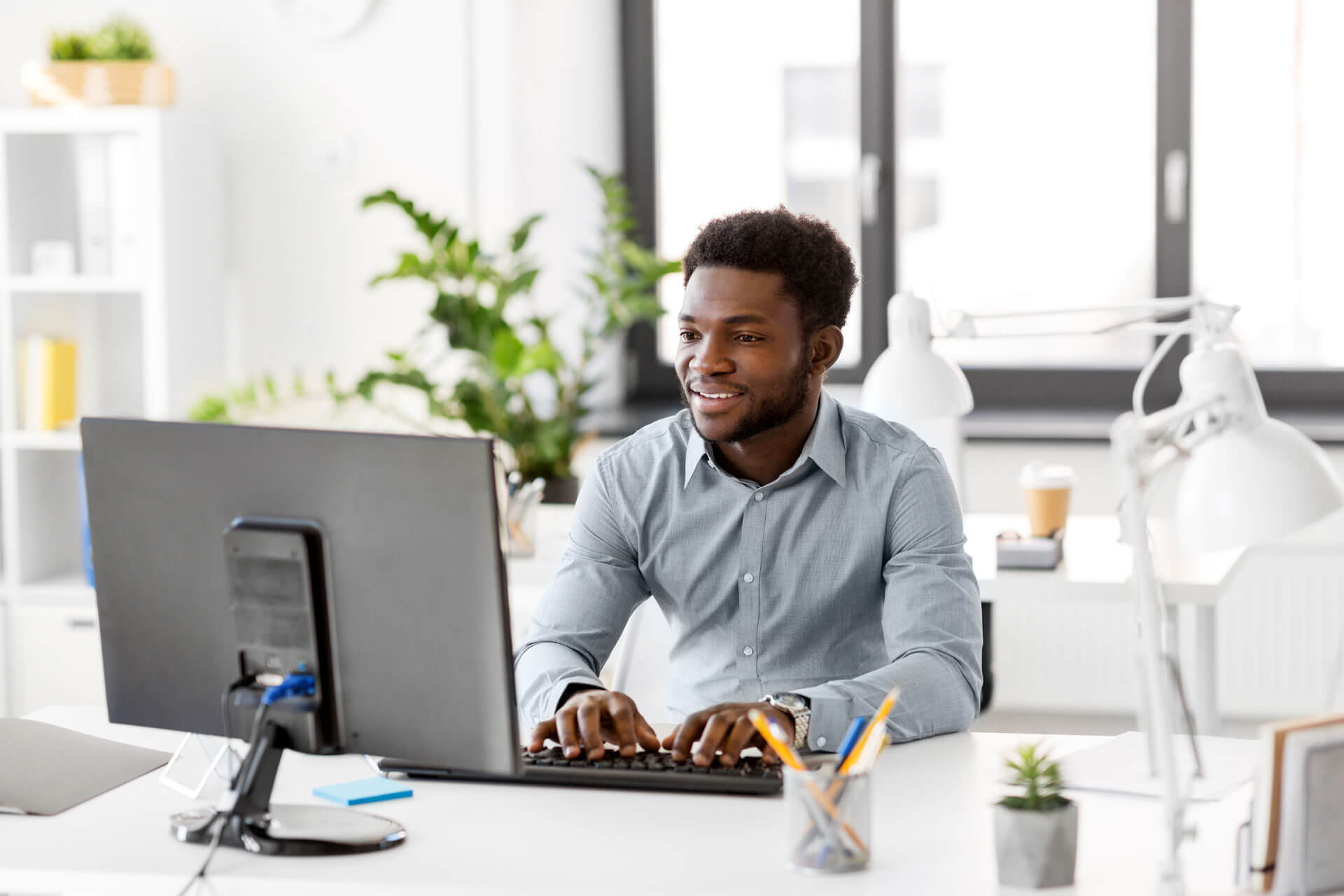 A man in a light gray shirt is sitting at a desk, typing on a keyboard and smiling at a computer screen. The office setting includes a large window, potted plants, a lamp, and organized stationery.