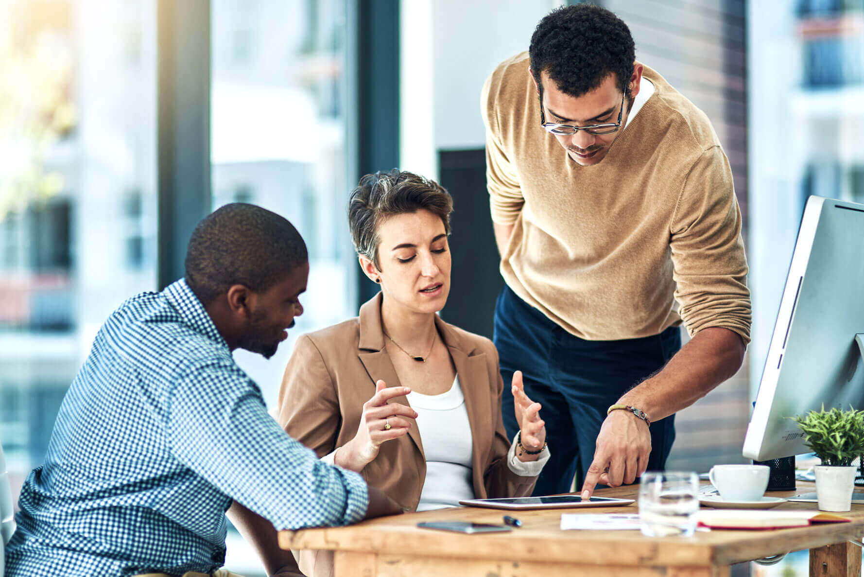 Three people are gathered around a table in an office setting. One person stands and points to documents on the table, while the other two sit and look intently at the paperwork. A computer monitor and glasses of water are also on the table.