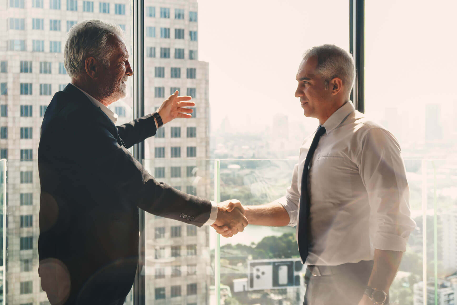 Two men shaking hands in a modern office with large windows, overlooking a cityscape. The man on the left, in a suit, gestures with his left hand, while the man on the right, in a dress shirt and tie, smiles. Natural light fills the room from outside.