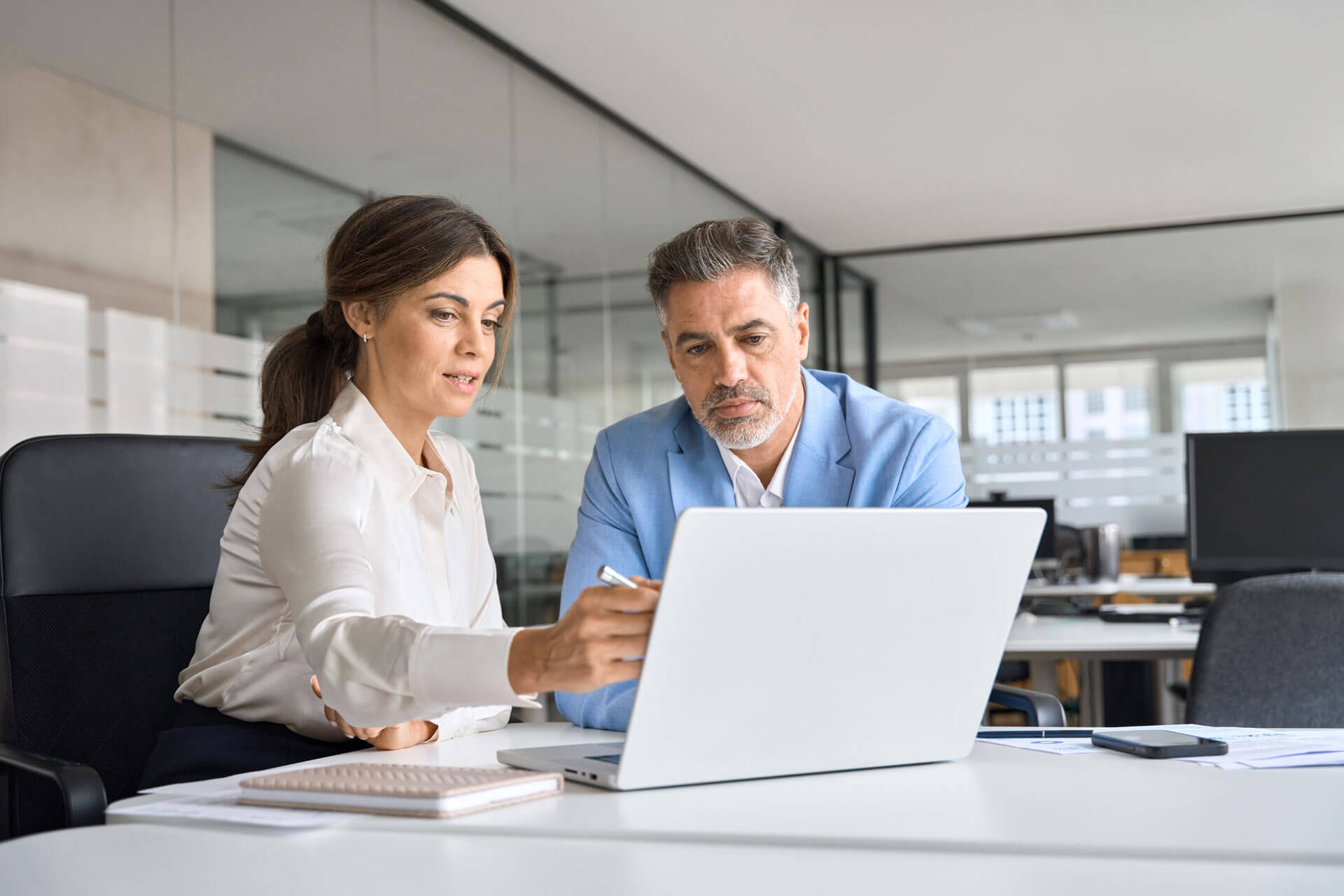 Two people sit at a desk in a modern office, looking at a laptop screen. The woman on the left points to the screen, while the man on the right, dressed in a light blue blazer, listens attentively. The background features glass walls and office equipment.