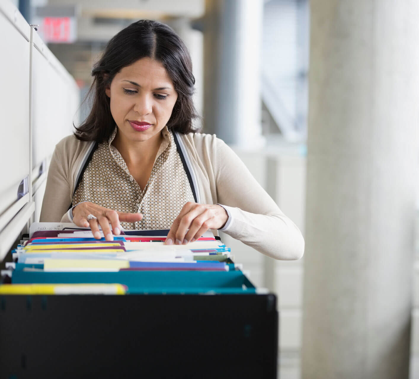 A woman in a beige blouse stands in an office, looking through colorful folders in an open filing cabinet. The background is blurred, showing a modern office setting with light gray and white tones.