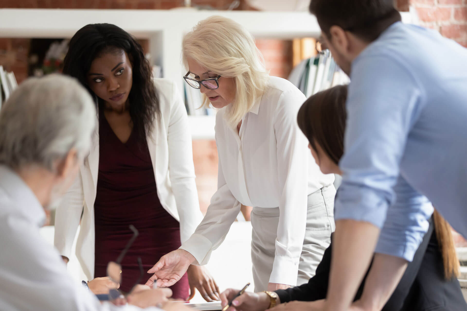 A diverse group of five people in a business meeting. A woman with glasses, standing and leaning over a table, discusses with colleagues who are intently listening and engaged. The setting appears to be an office with books and documents in the background.