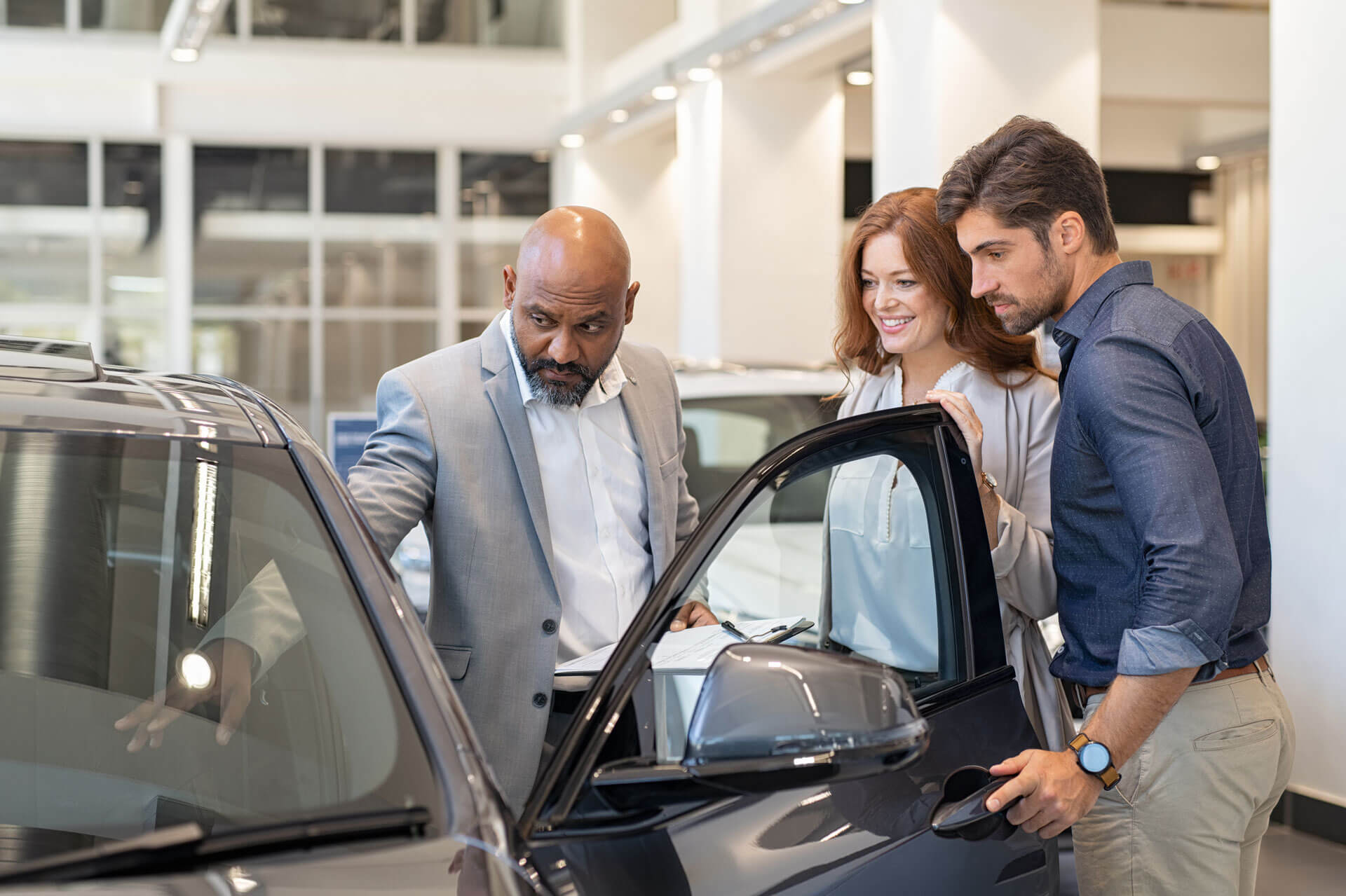 A man wearing a gray suit shows a car to a couple at a dealership. The couple, standing together, looks at the car interior while the woman smiles. The environment appears bright and modern with cars visible in the background.
