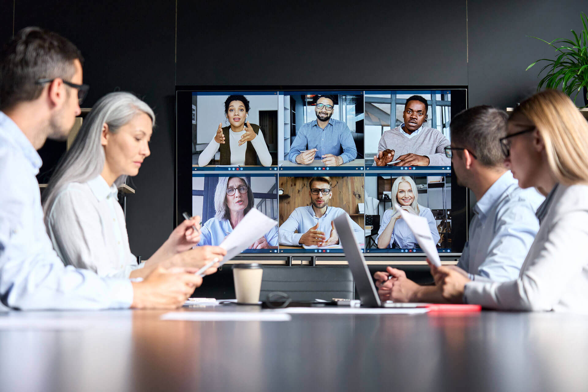 A group of four coworkers sits around a conference table, each with papers and laptops, participating in a video conference. On the screen in front of them are eight other participants, each in their own video window, engaging in the virtual meeting.
