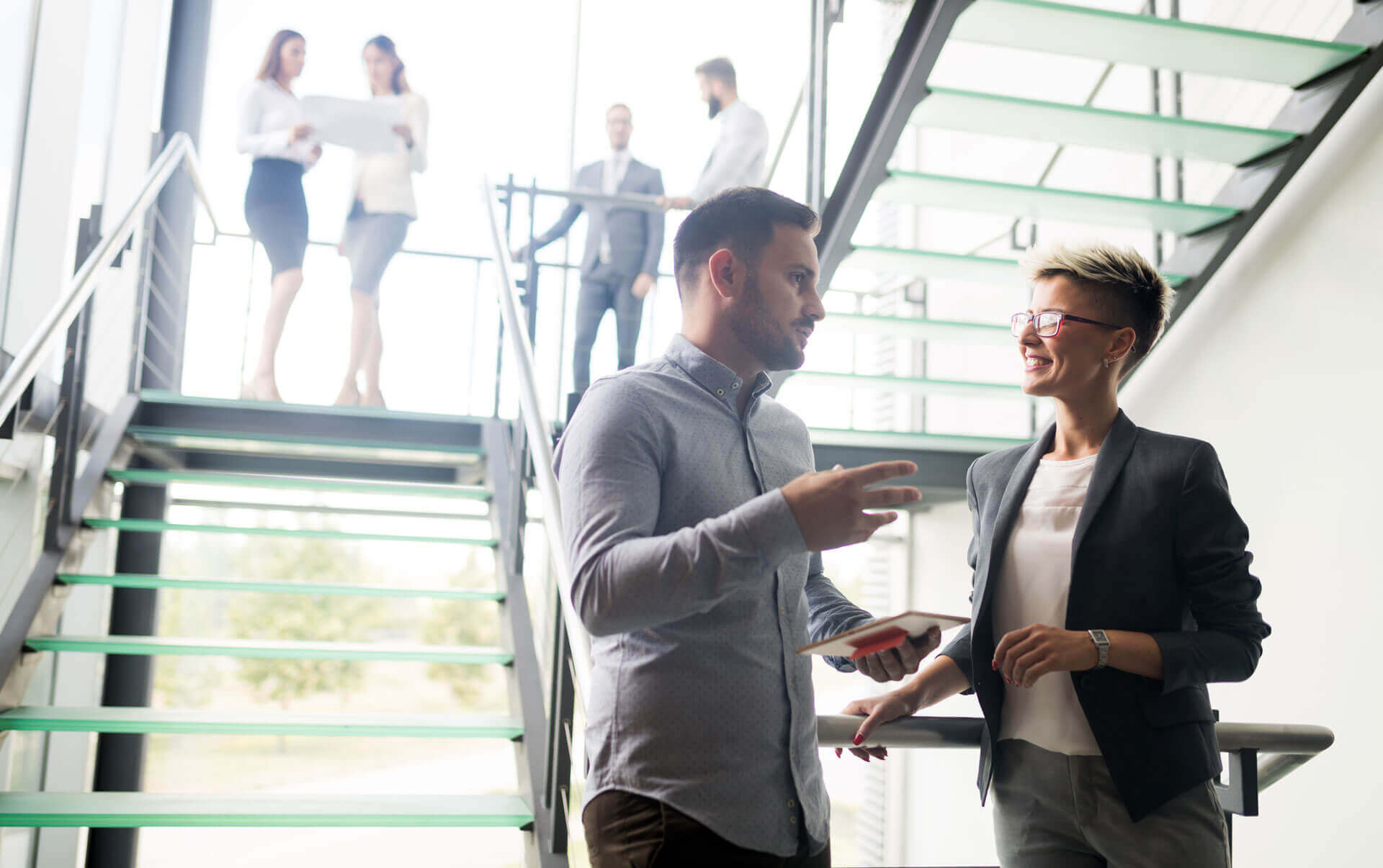 Two colleagues are having a conversation beside a staircase in a modern office space. One is a man holding a tablet, the other is a woman with short hair and glasses. In the background, four other professionals are engaged in discussions on the stairs and landing.