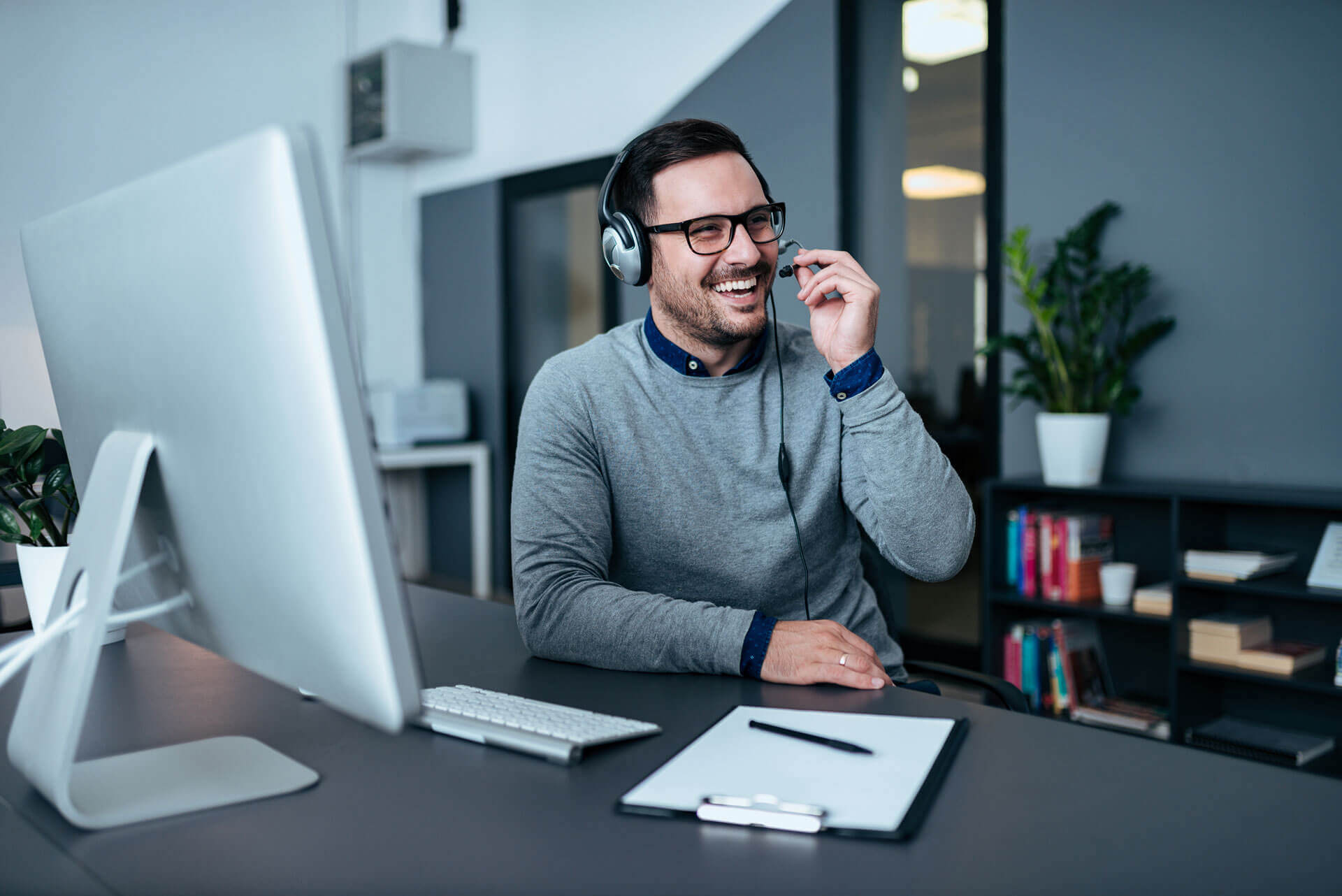 A man with glasses and headphones is sitting at a desk in a modern office. He is smiling and appears to be engaged in a conversation. On the desk are an iMac, keyboard, mouse, notepad, and pen. There is a plant in the background and books on a shelf.