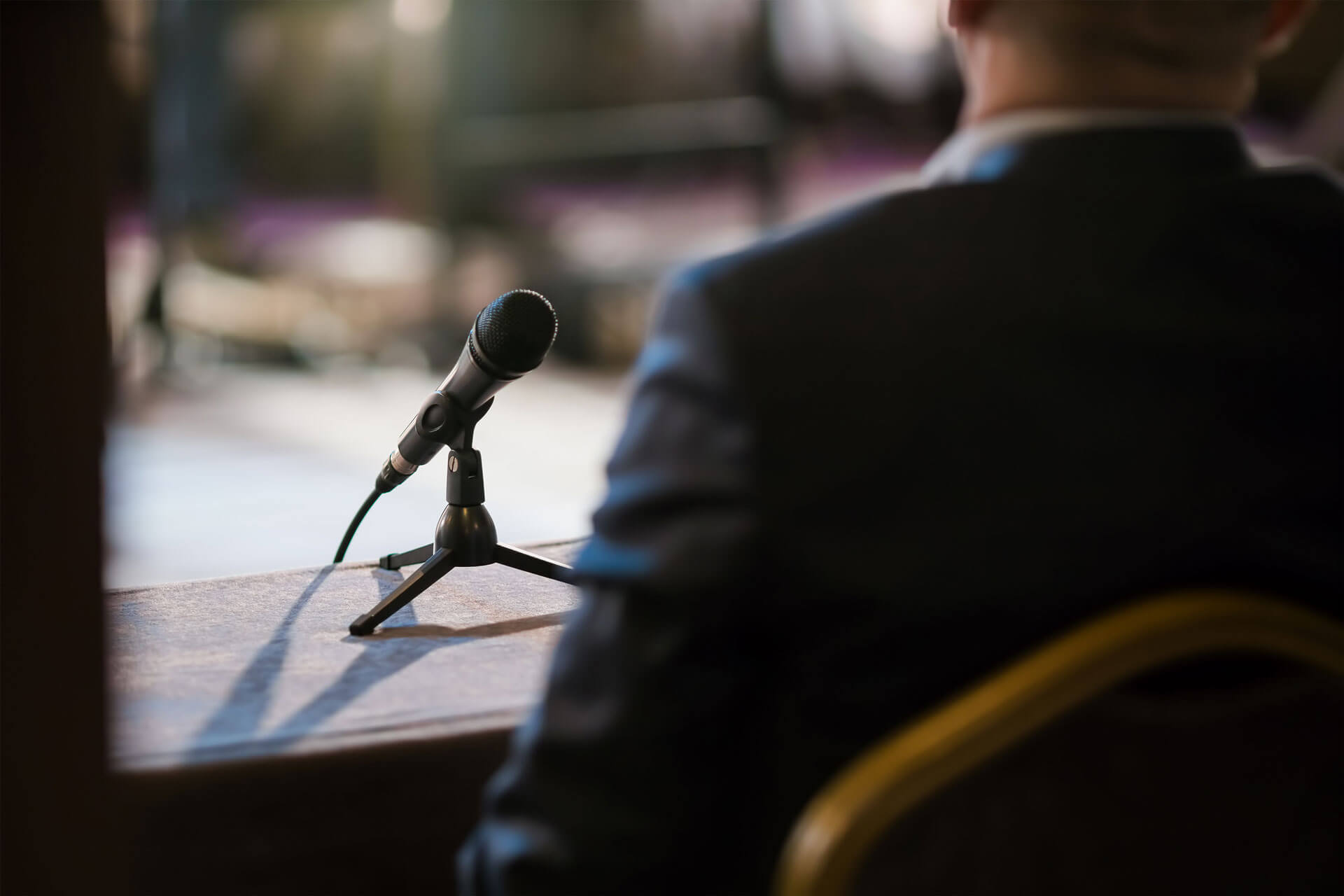 A person in a suit sits at a table with a microphone on a stand, facing away from the camera. The setting appears to be a conference or meeting room. The background is blurred.