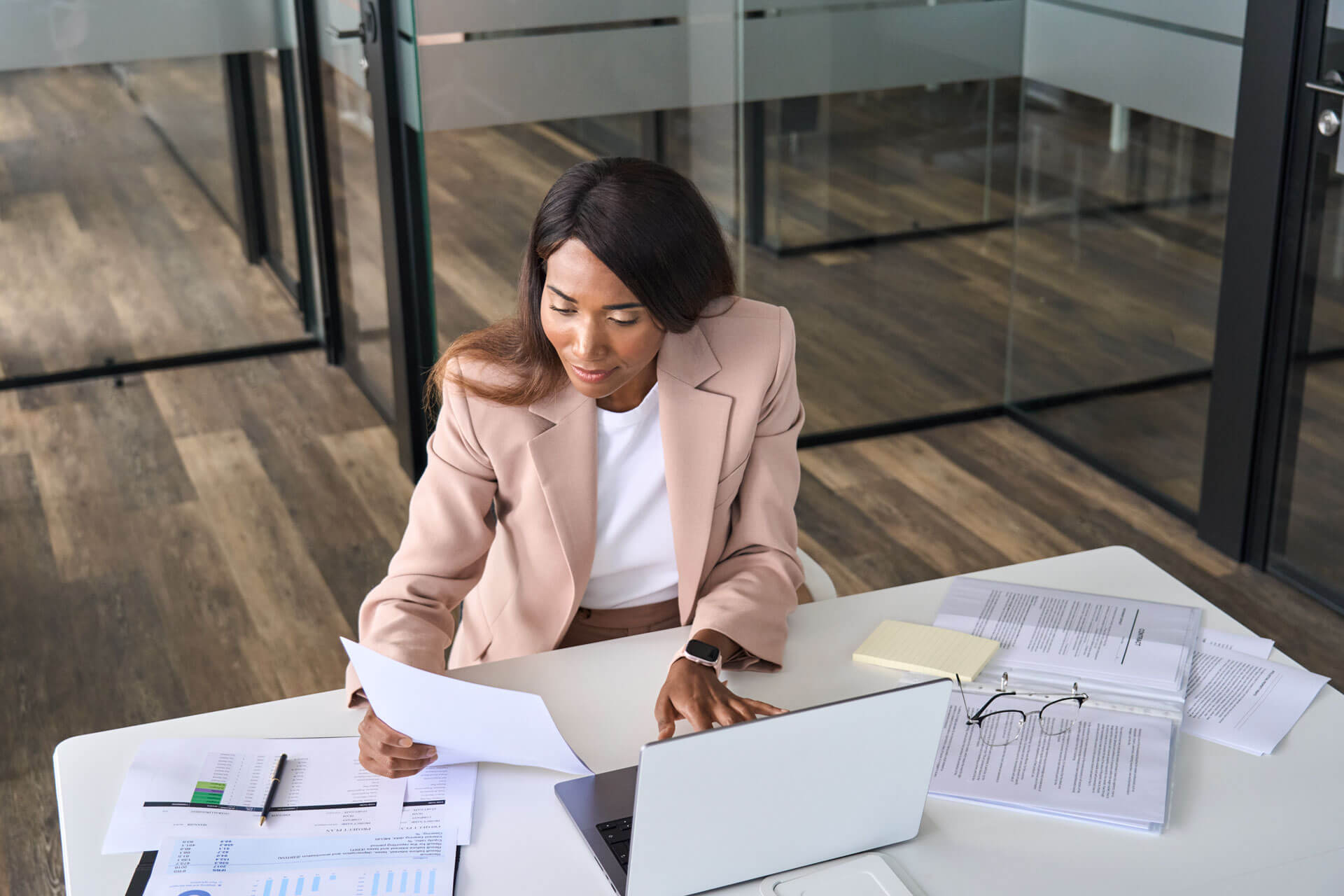A woman wearing a beige blazer sits at a desk in a modern office, reviewing documents. She has a laptop open in front of her, along with papers, a notebook, and a pair of glasses on the table. Glass walls and a wooden floor are visible in the background.