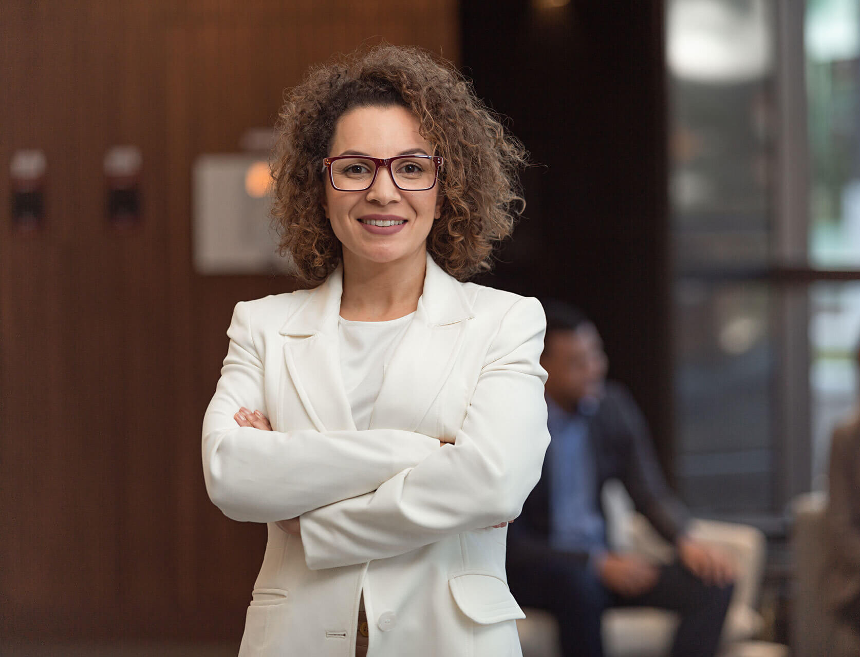 A woman with curly hair and glasses is smiling and standing with her arms crossed. She is wearing a white blazer and a white blouse. In the background, two people are seated and conversing in a modern, well-lit room with large windows.