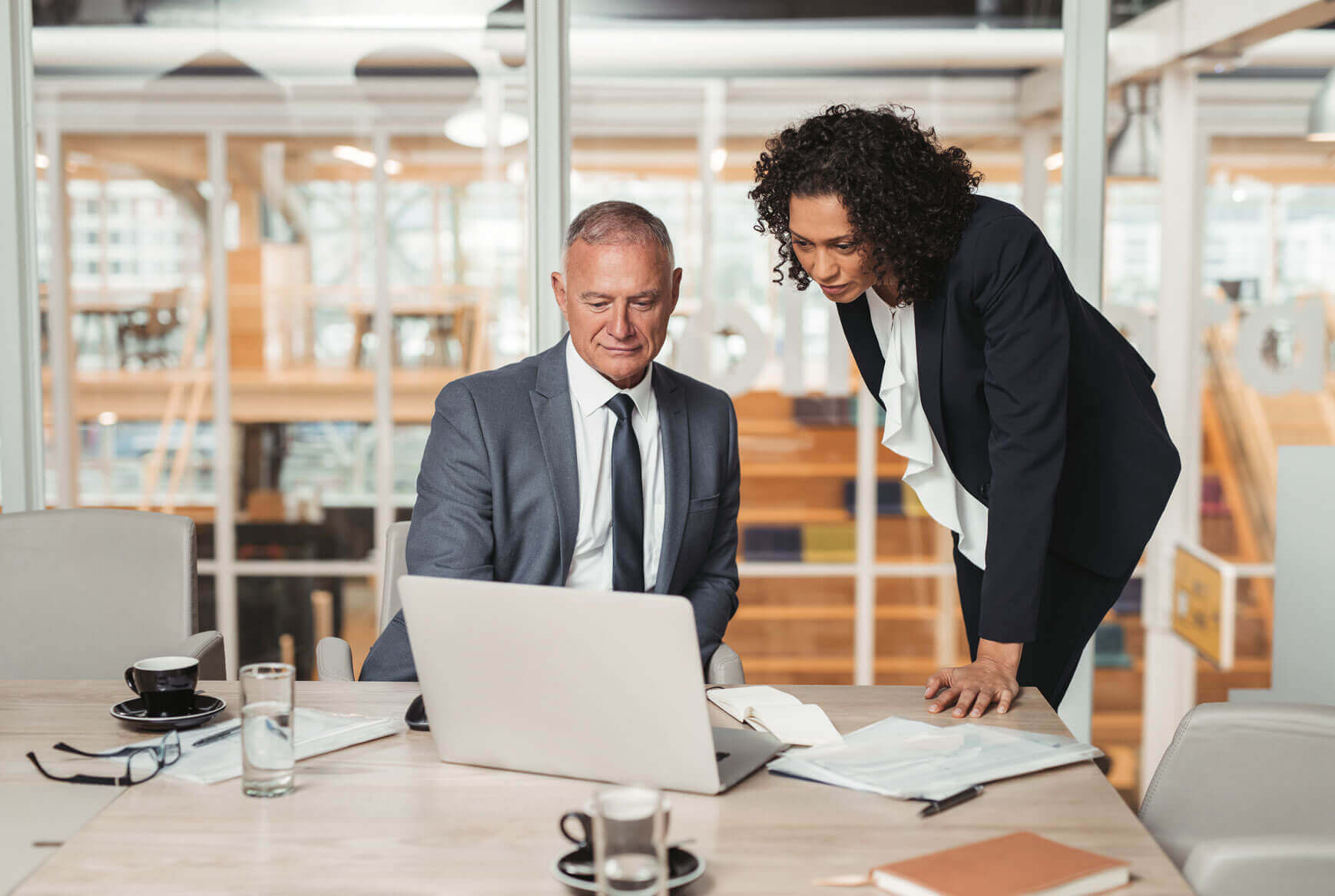 Two business professionals are in a modern office. A man sits at a table with a laptop, while a woman stands next to him, looking at the screen. The table has a notebook, documents, and coffee cups. Glass walls and an open office space are in the background.