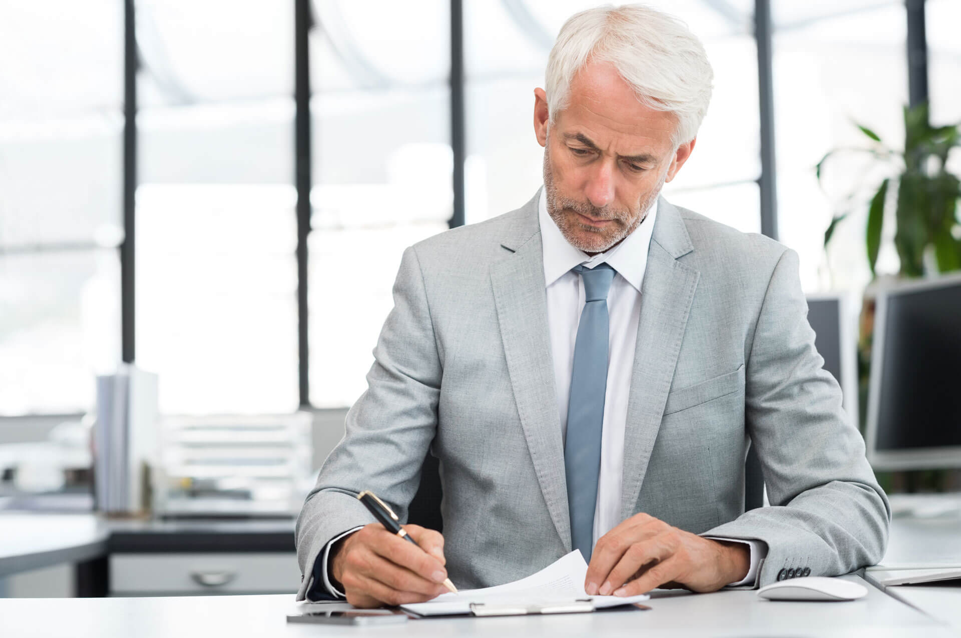 An older man with white hair and a beard, wearing a light grey suit and blue tie, is seated at a desk in a modern office. He is focused on writing in a notebook with a pen. Large windows in the background let in natural light.