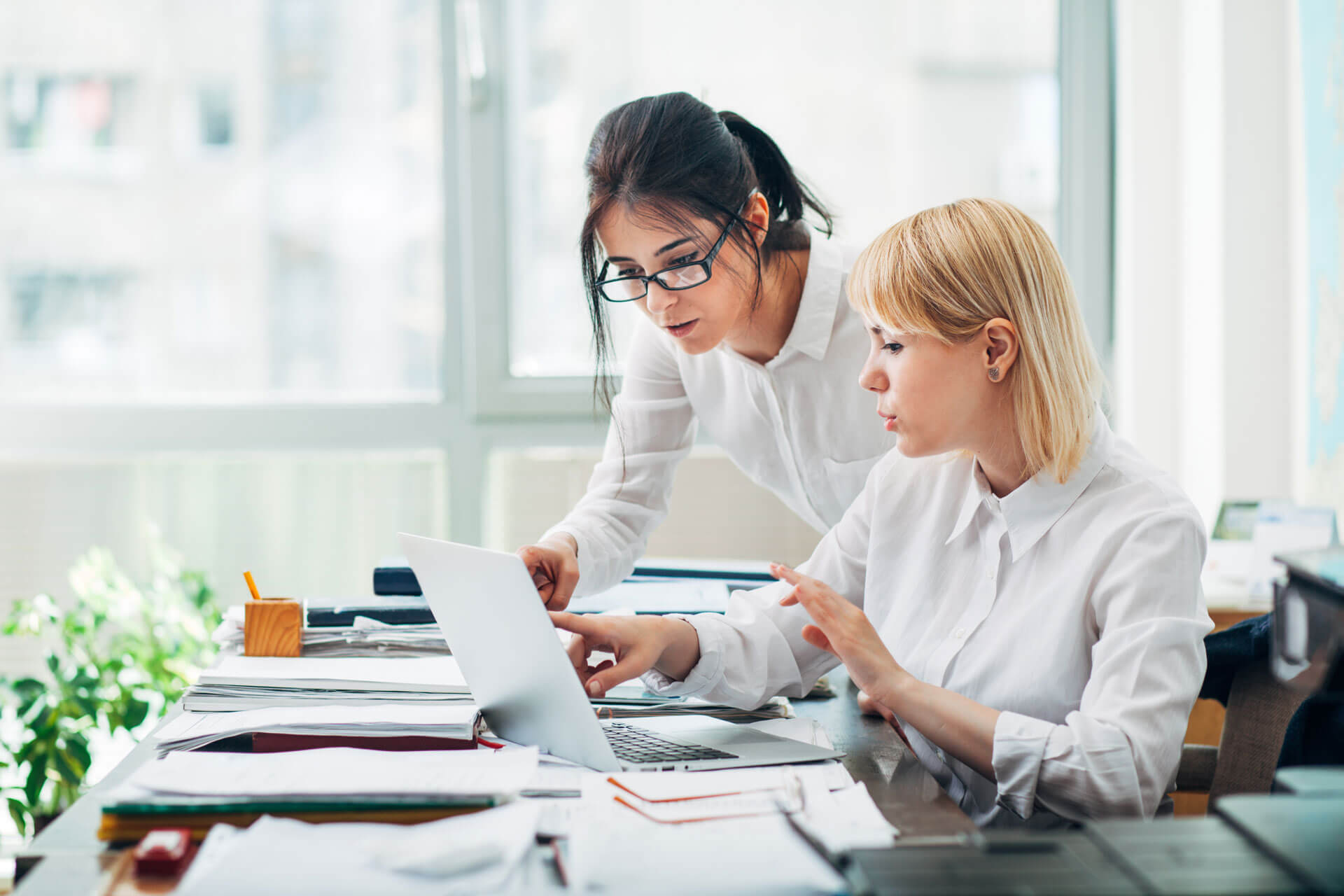 Two women in an office are collaborating. One, with glasses and dark hair, stands, leaning over to point at the laptop screen. The other, with blonde hair, is seated, attentively looking at the screen. The desk is cluttered with papers and office supplies.