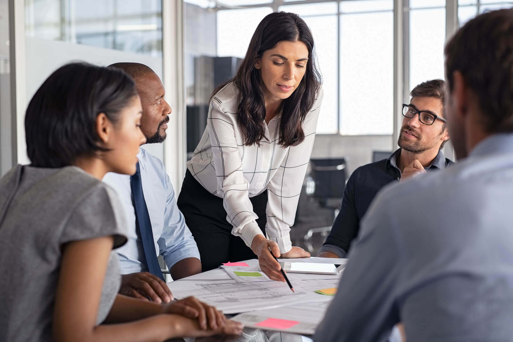 A woman in a white blouse stands and points to a document on a table, representing Cogency Global's document retrieval services. Four colleagues, two men and two women, sit around the table, attentively listening.