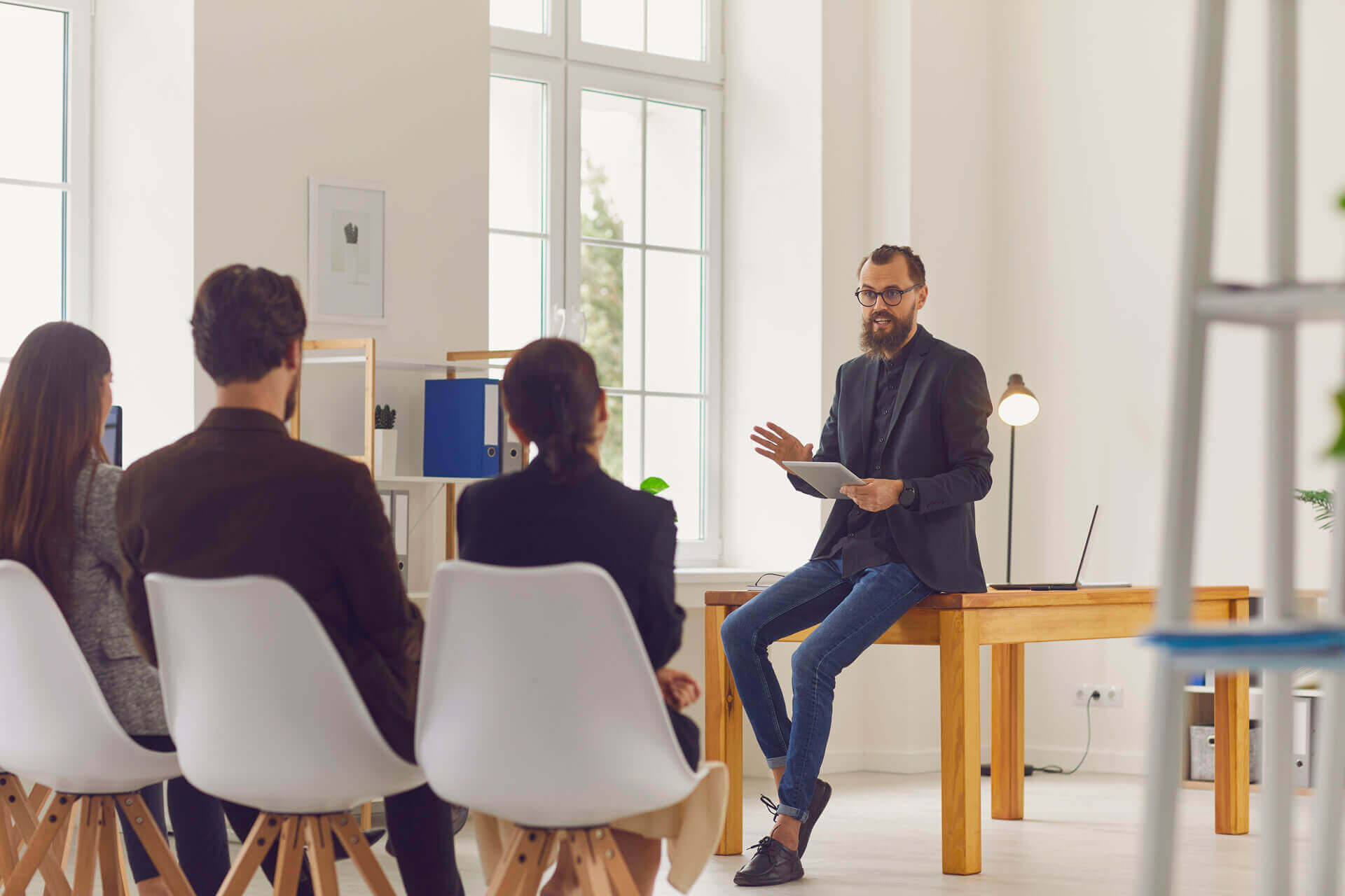 A bearded man wearing glasses and a black blazer sits on a desk, holding a tablet, while speaking to three seated people in a bright, modern office space. The listeners are in casual attire and facing him attentively.