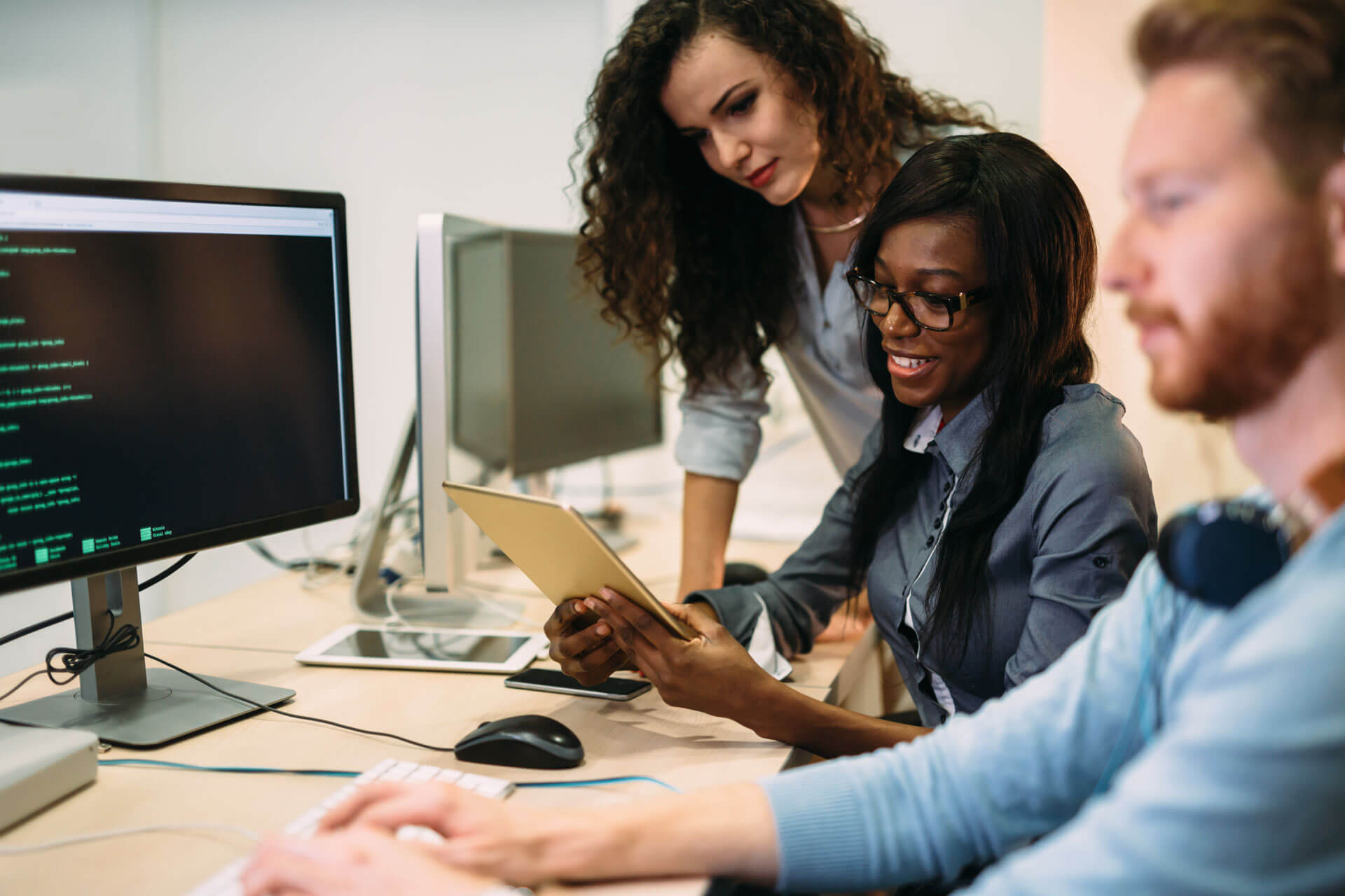 Three people collaborate in an office setting. A man types on a computer, displaying code. A woman holds a tablet, and another woman observes. They all focus on the computer screen. Desks and computer equipment are visible in the background.