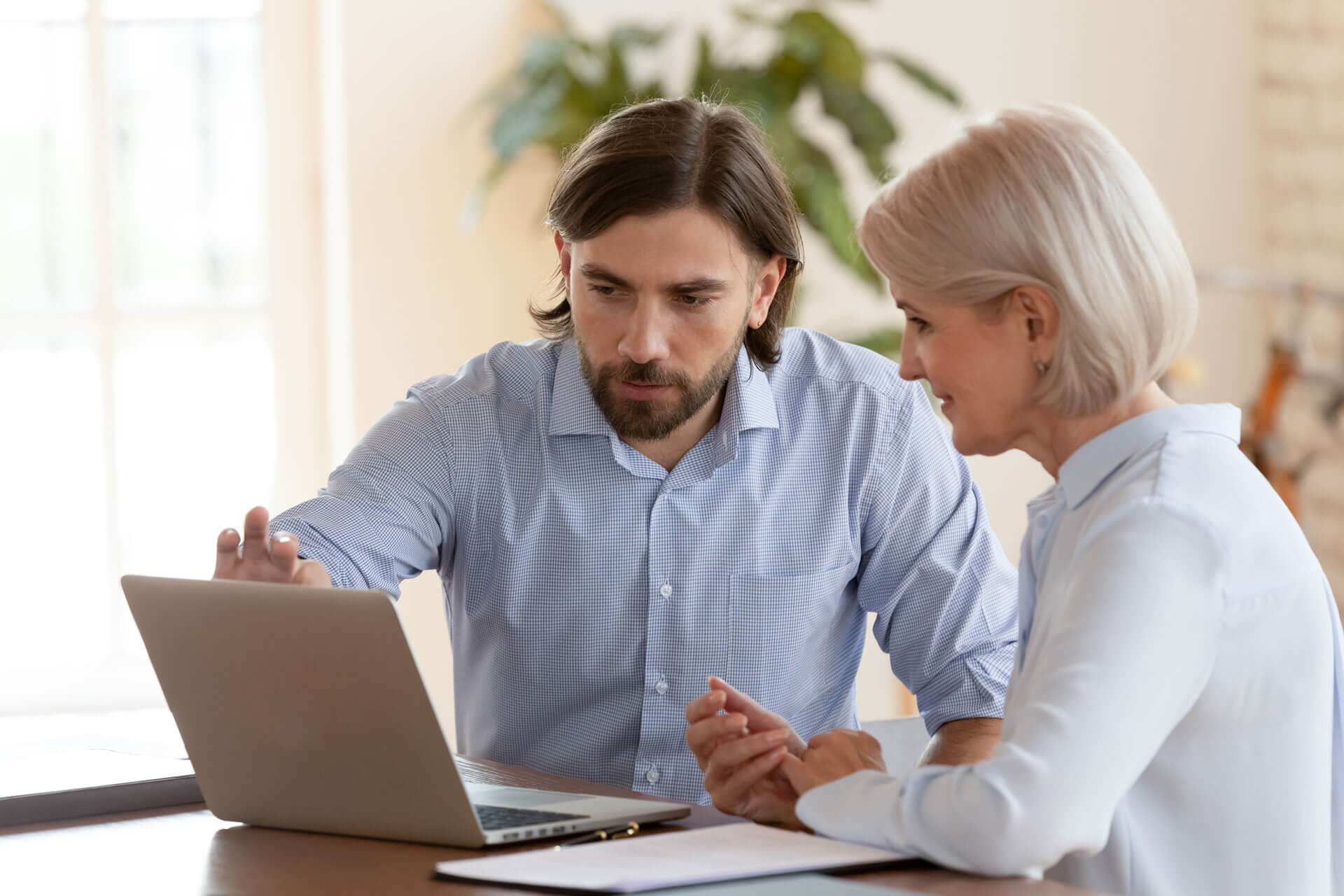 A man with medium-length dark hair and a beard is sitting and pointing at a laptop screen. A woman with short blonde hair sits next to him, attentively looking at the screen. Both are dressed in business casual attire, and a plant is visible in the background.