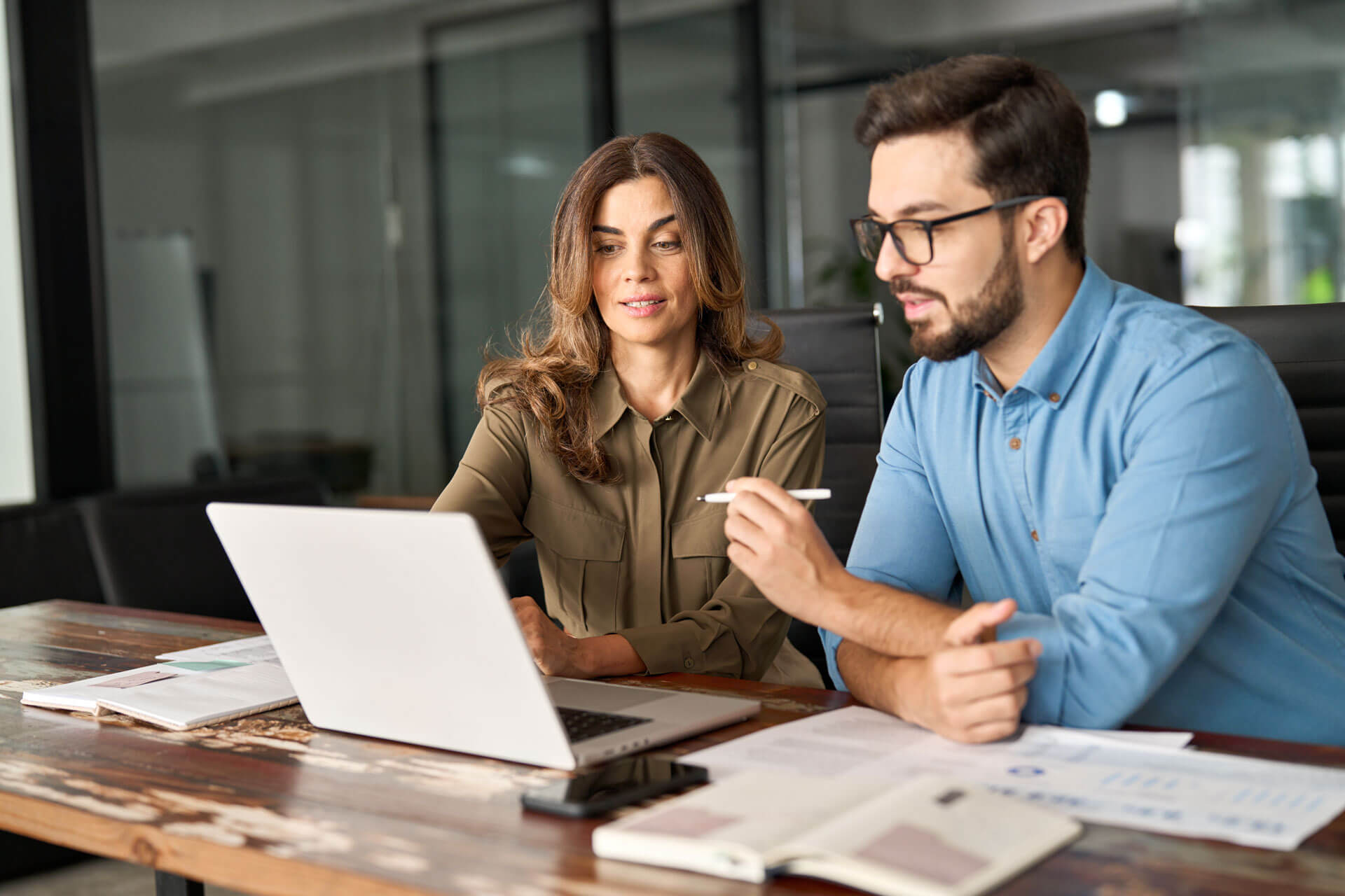 Two people are seated at a wooden table in an office, looking at a laptop. The person on the left, with long hair, is in an olive shirt. The person on the right, with glasses and a beard, is in a blue shirt, holding a pen. Papers are scattered on the table.