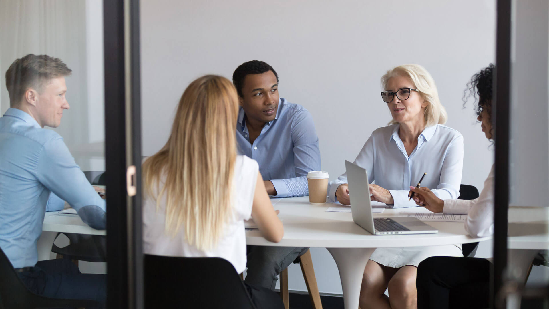 A diverse group of people sit around a table having a meeting. There are three women and two men. A laptop and coffee cup are on the table. The setting is a modern office with a white wall background.