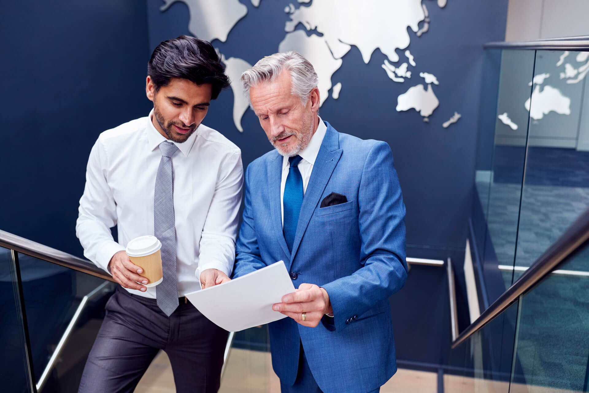 Two men in business attire stand on a staircase, reviewing documents. One holds a coffee cup. A world map decor is visible on the dark wall behind them. The setting appears to be an office or professional environment.