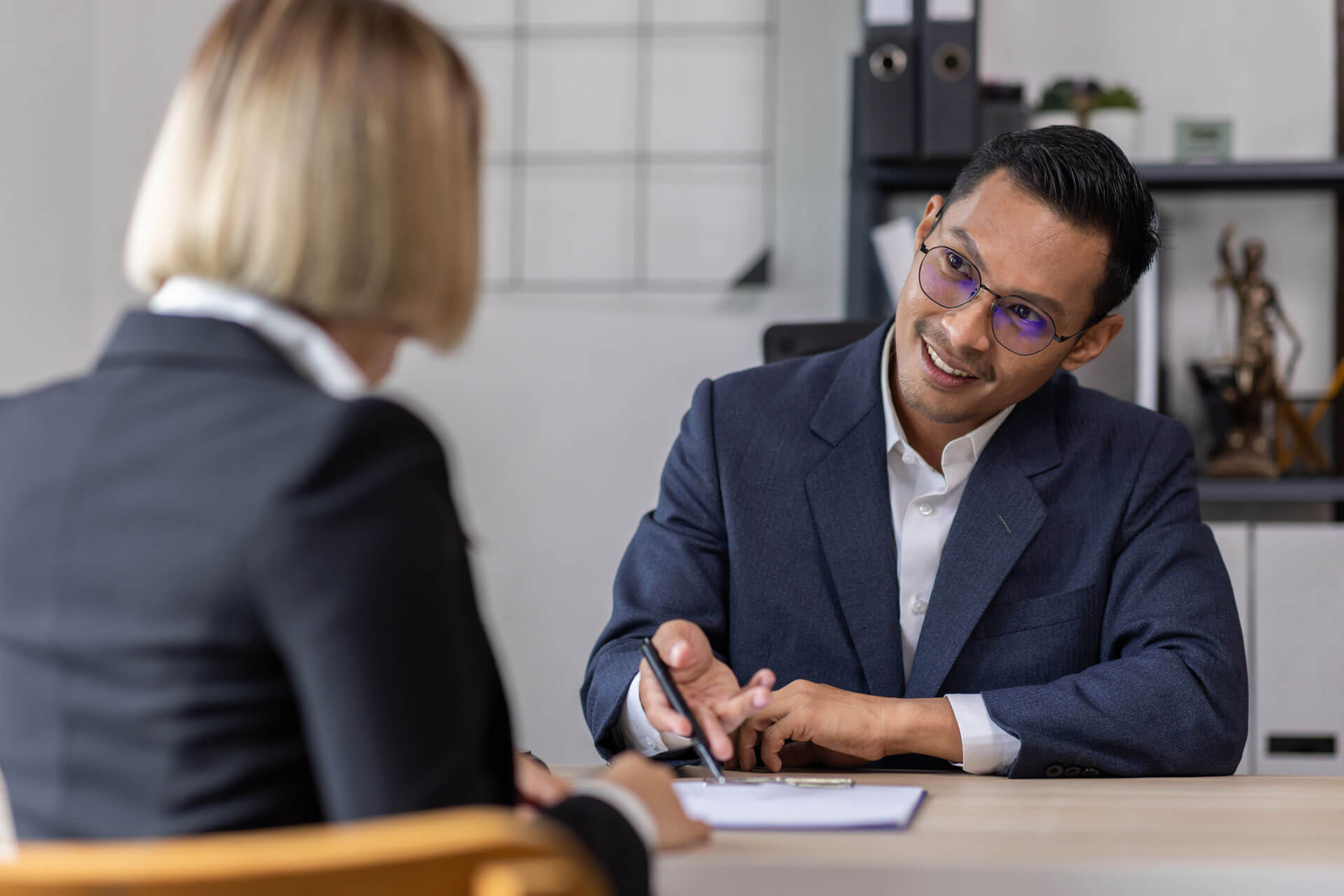 A man in a suit with glasses sits at a desk holding a pen and looking at a woman across from him. The woman, with short blonde hair, is wearing a black blazer. They appear to be having a professional conversation. Office supplies are visible in the background.