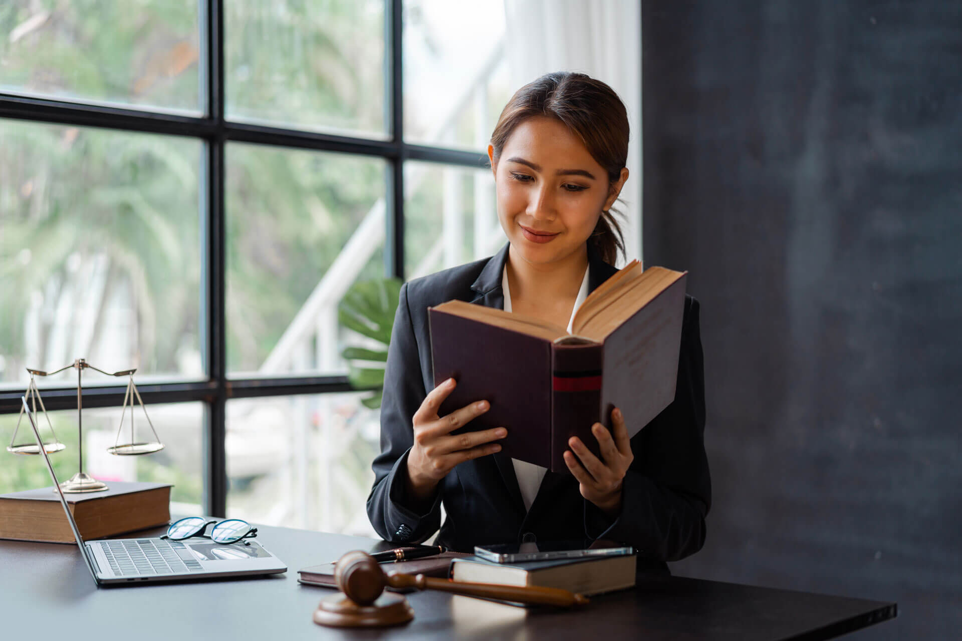 A woman in professional attire sits at a desk reading a book. The desk has a laptop, a gavel, and scales of justice. Behind her is a large window letting in natural light. The scene is set in an office with a dark wall.