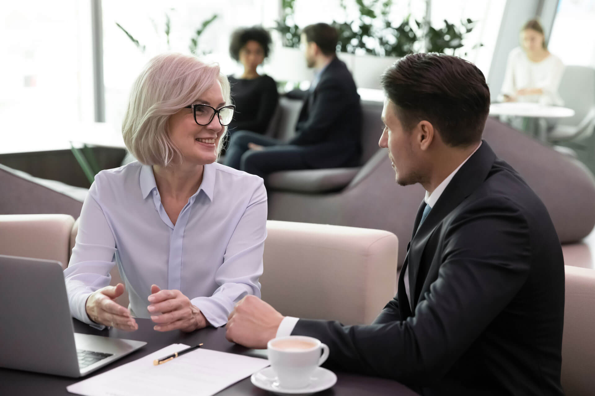 A woman with short blonde hair and glasses is smiling and discussing something with a man in a suit, seated across from her in a modern office setting. A laptop, paper, and coffee cup are on the table. In the background, people are engaged in conversations.