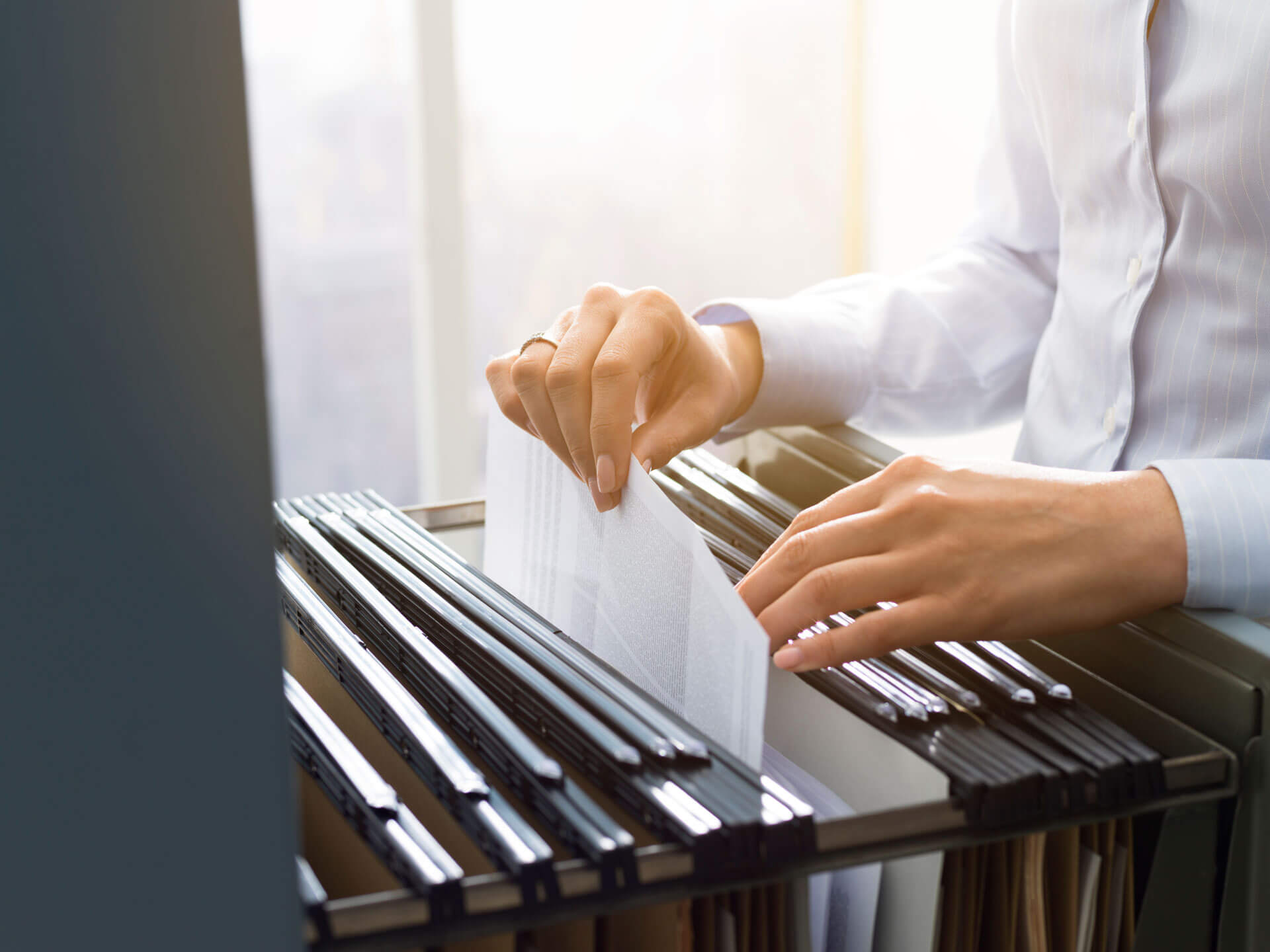 A person wearing a white shirt is sorting through hanging file folders in a filing cabinet. The person is pulling out a document from one of the folders. The background is blurred, suggesting an office environment with natural light coming through the window.