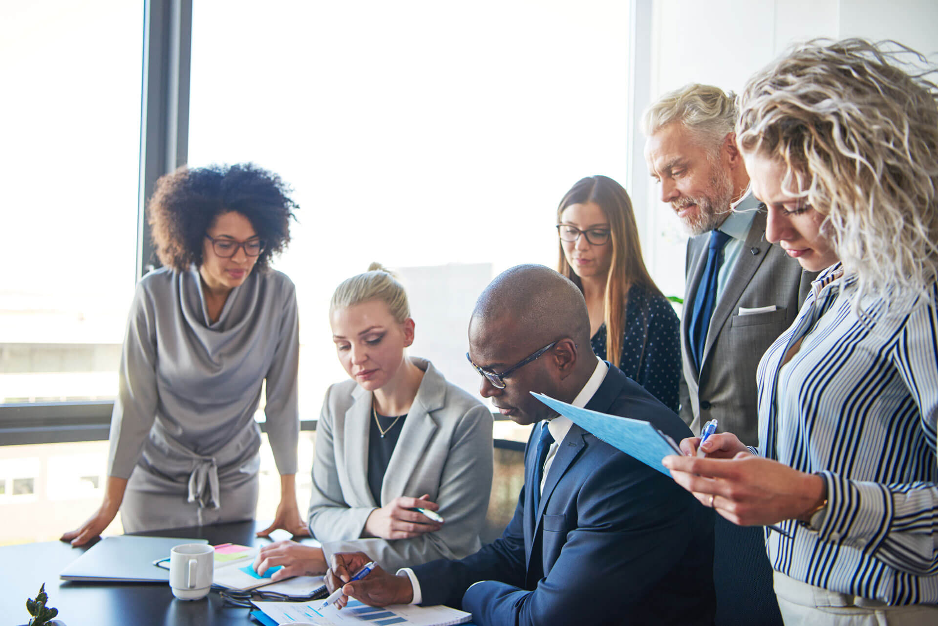 A group of five business professionals of diverse backgrounds are gathered around a table in a bright office. Two women are standing on the left, while a man in a dark suit is seated, writing. Another woman and man are standing, closely observing the documents.