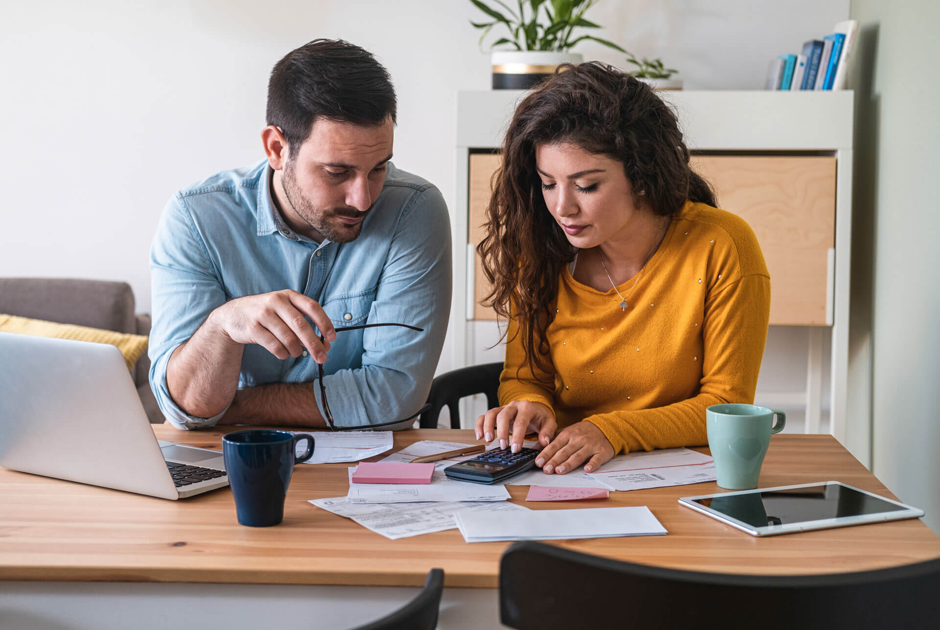 A man and woman sit at a wooden table working on their finances. The man holds a pen and appears to be explaining something, while the woman uses a calculator. The table is cluttered with documents, a laptop, two coffee mugs, and a tablet.