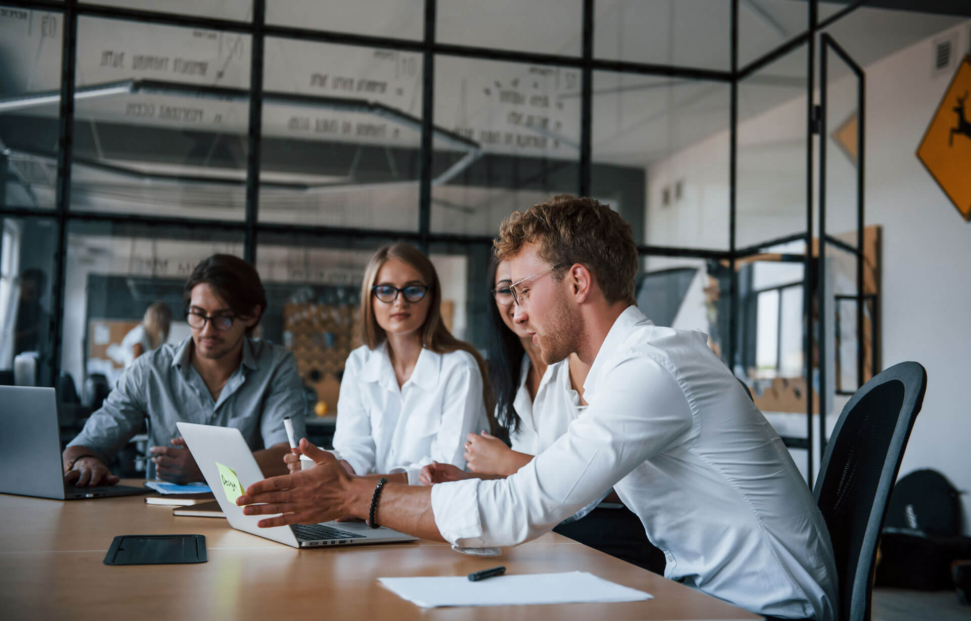A group of four people in a modern office room, sitting at a table with laptops and papers. They are engaged in a nonprofit formation discussion, with one person pointing at a laptop screen. The room has large windows and glass partitions.