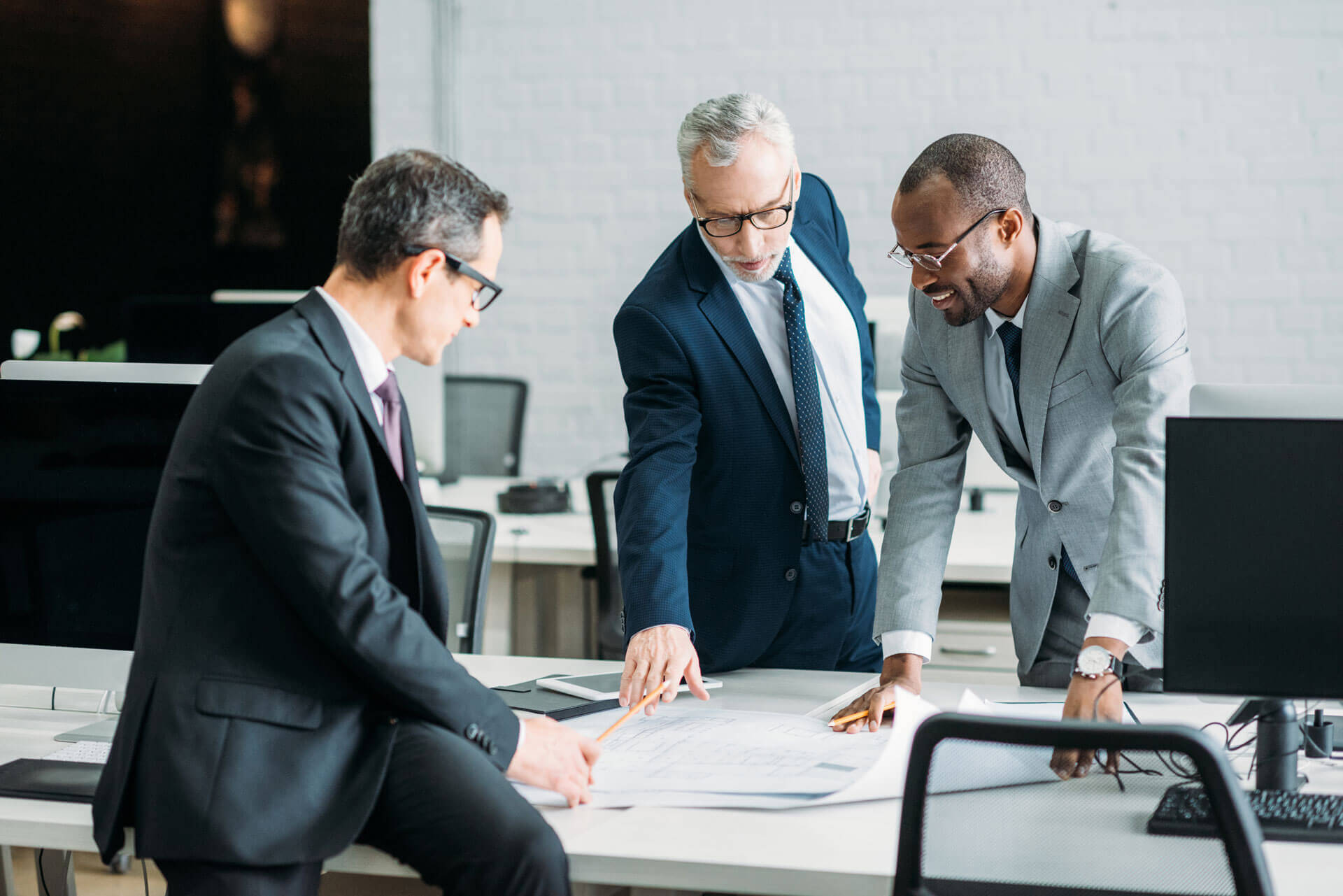 Three businessmen in suits collaborate over a large document on a desk in a modern office, representing clients who may need Cogency Global’s process agent services. Two men are standing, one pointing to a detail on the paper, while the third man, sitting on the desk, looks on attentively.
