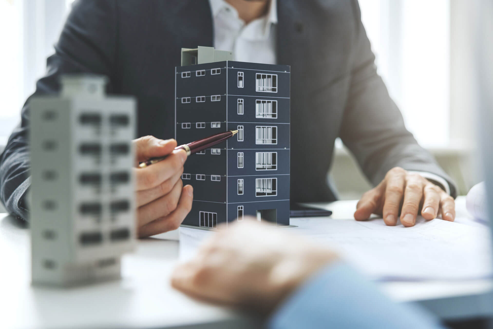 Two people in business attire are sitting at a table with architectural models of buildings. One person is pointing at a model with a pencil, and papers are scattered on the table, suggesting a discussion about construction or real estate.