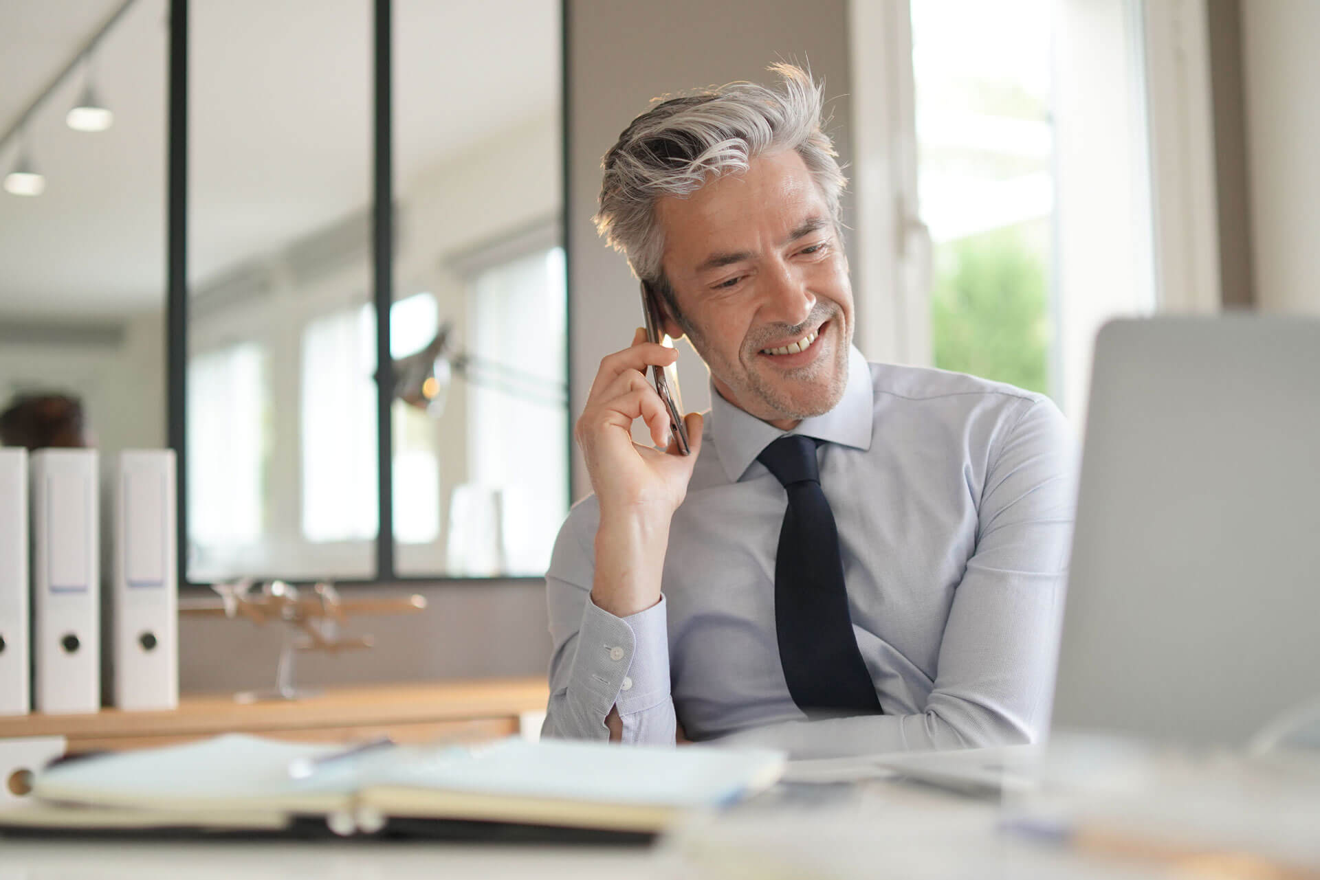 Smiling man with gray hair in a blue shirt and tie talks on a smartphone while looking at a laptop in a bright office. Papers and folders are on the desk, and a window is in the background.